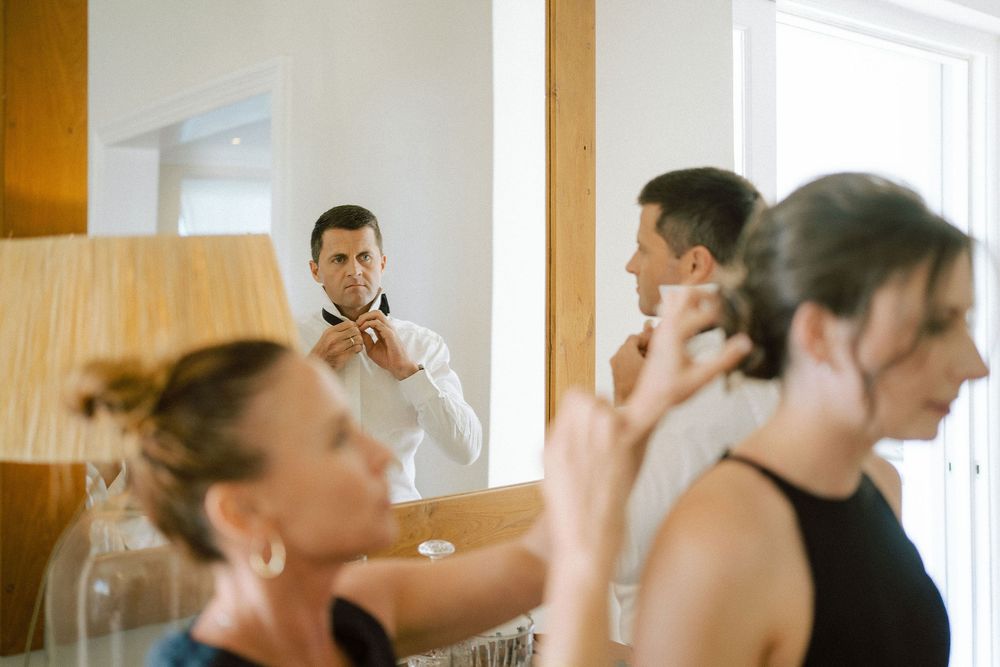 Groom in a white shirt adjusts his bow tie in a mirror while two women in the foreground get ready for a wedding.