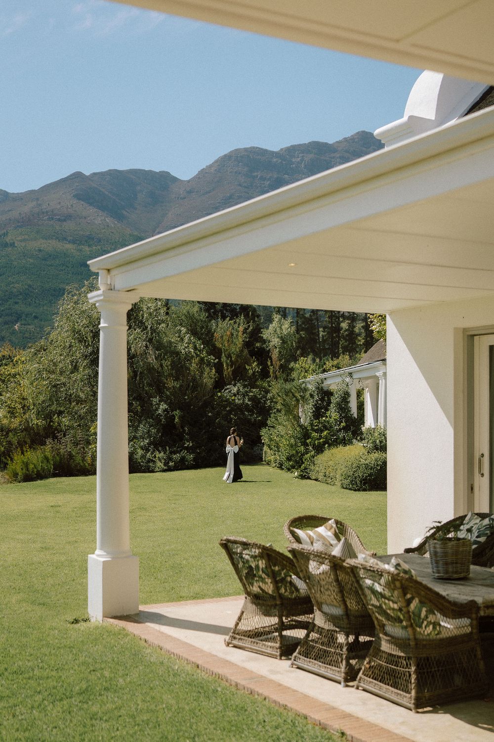 Covered veranda with wicker dining chairs overlooks a sunny lawn where a couple embraces, with lush trees and mountains in the background.