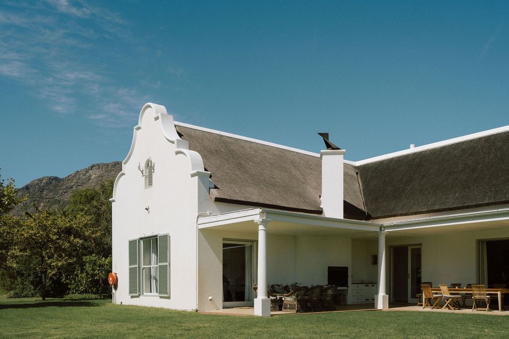 White Cape Dutch style house with curved gable, thatched roof and covered veranda, set on a green lawn with mountains and blue sky behind.