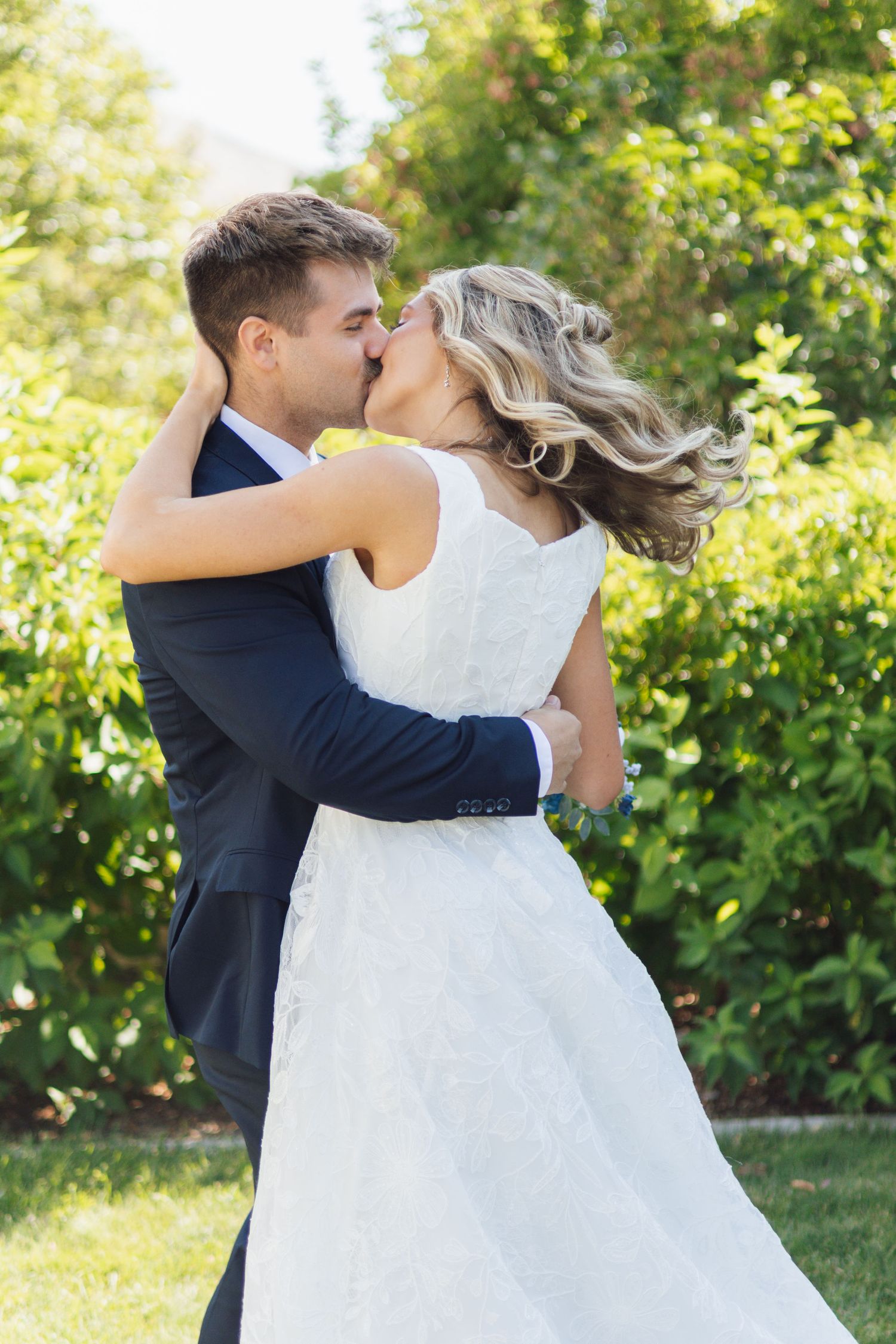 A bride in a white lace gown and a groom in a navy suit share a kiss in a sunny garden surrounded by green hedges.