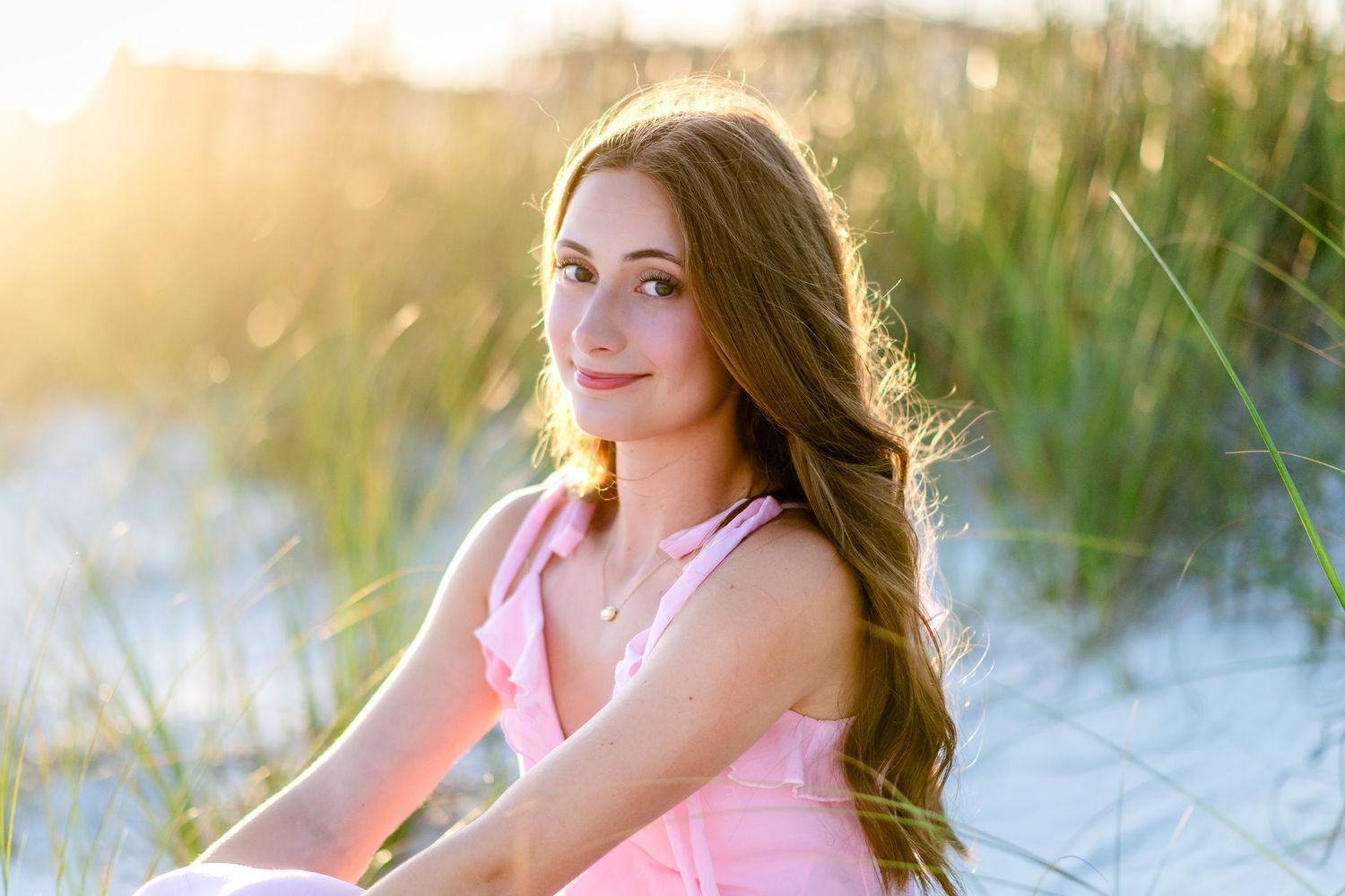 Young woman with long brown hair in a pink tank top sits among beach dune grass, smiling softly in warm golden sunset light.