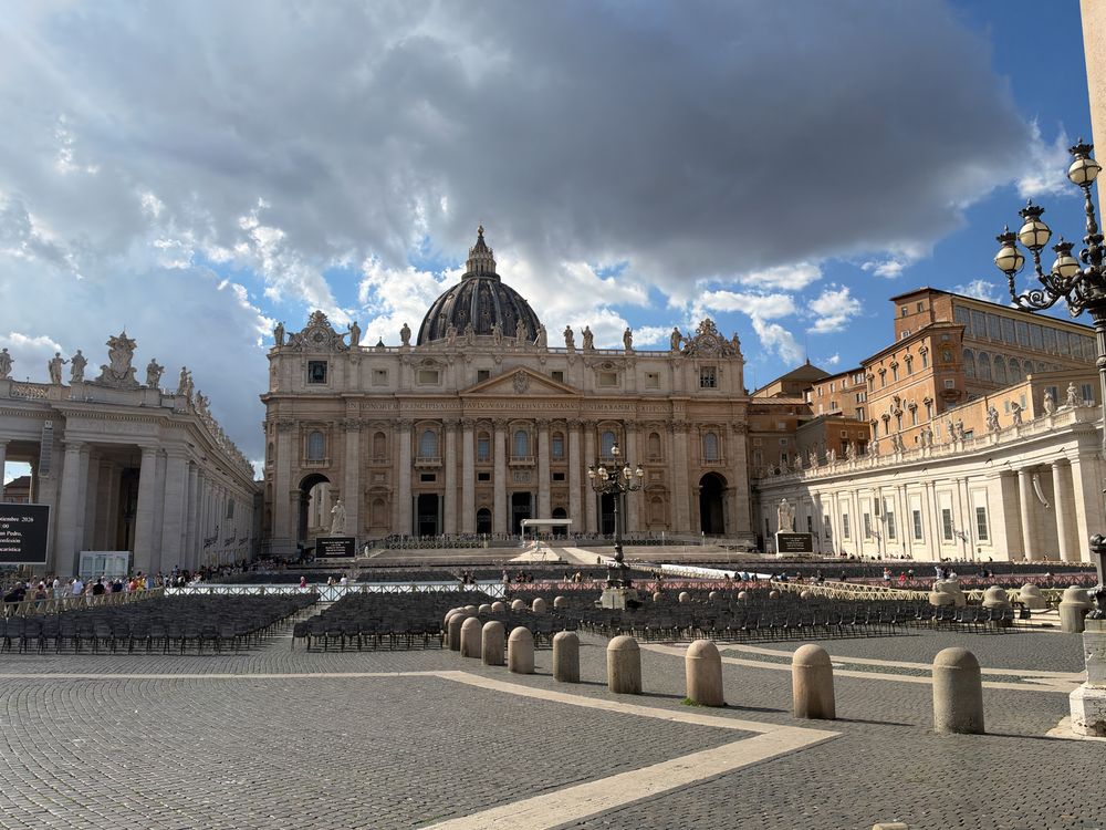 St. Peter's Basilica and the colonnaded St. Peter's Square in Vatican City under a dramatic cloudy blue sky with empty chairs set out.