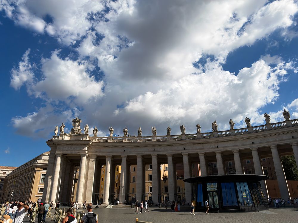 Tourists gather beneath a dramatic cloudy blue sky in front of the grand colonnade topped with statues in St. Peter's Square, Vatican City.