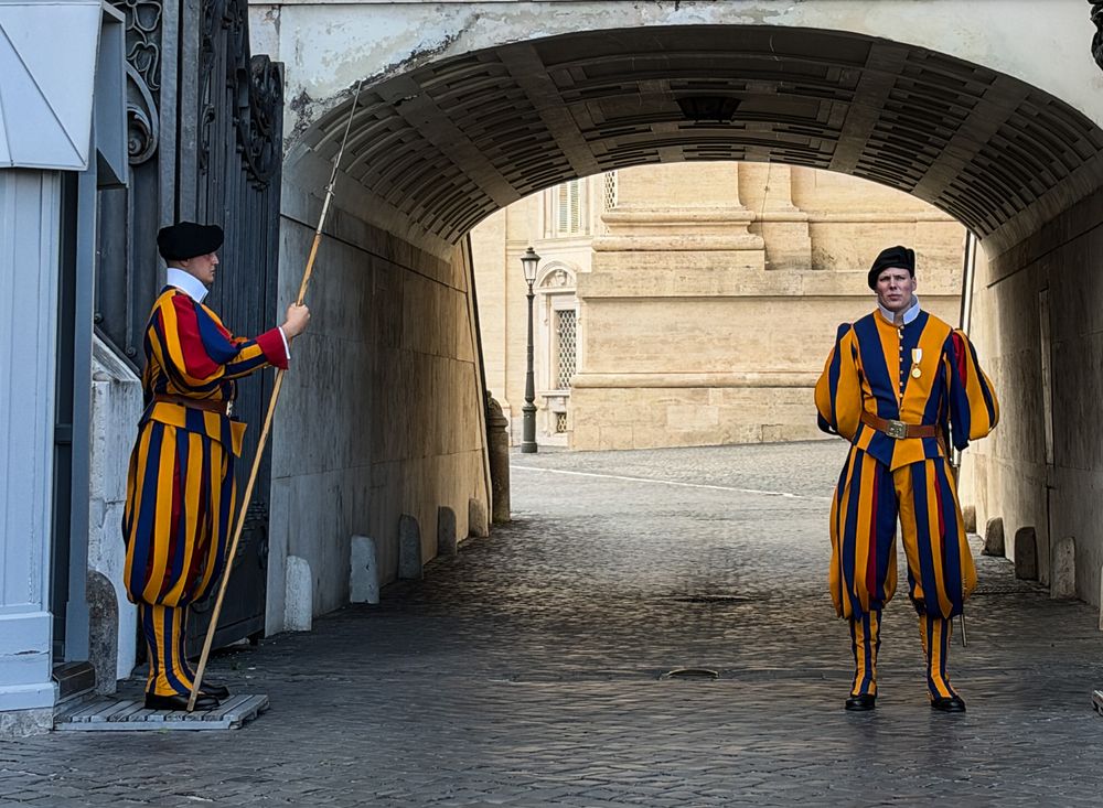 Two Swiss Guards in striped blue, red and yellow uniforms stand at attention beneath a stone archway at Vatican City.