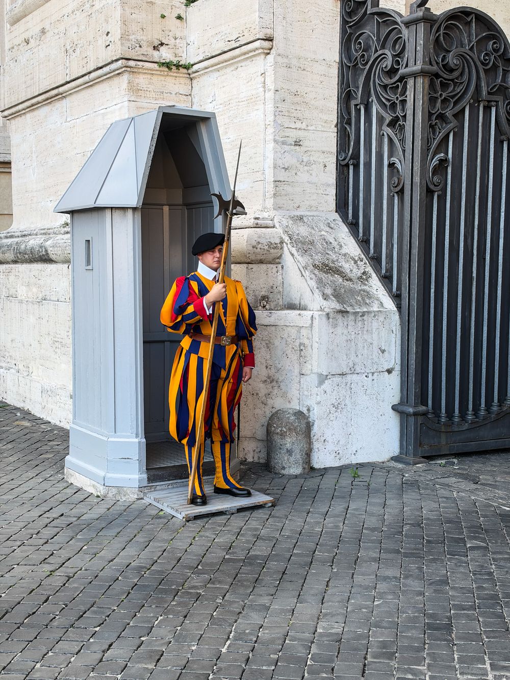 Swiss Guard in a striped blue, red and yellow Renaissance uniform holding a halberd beside a gray sentry box and ornate iron gate.