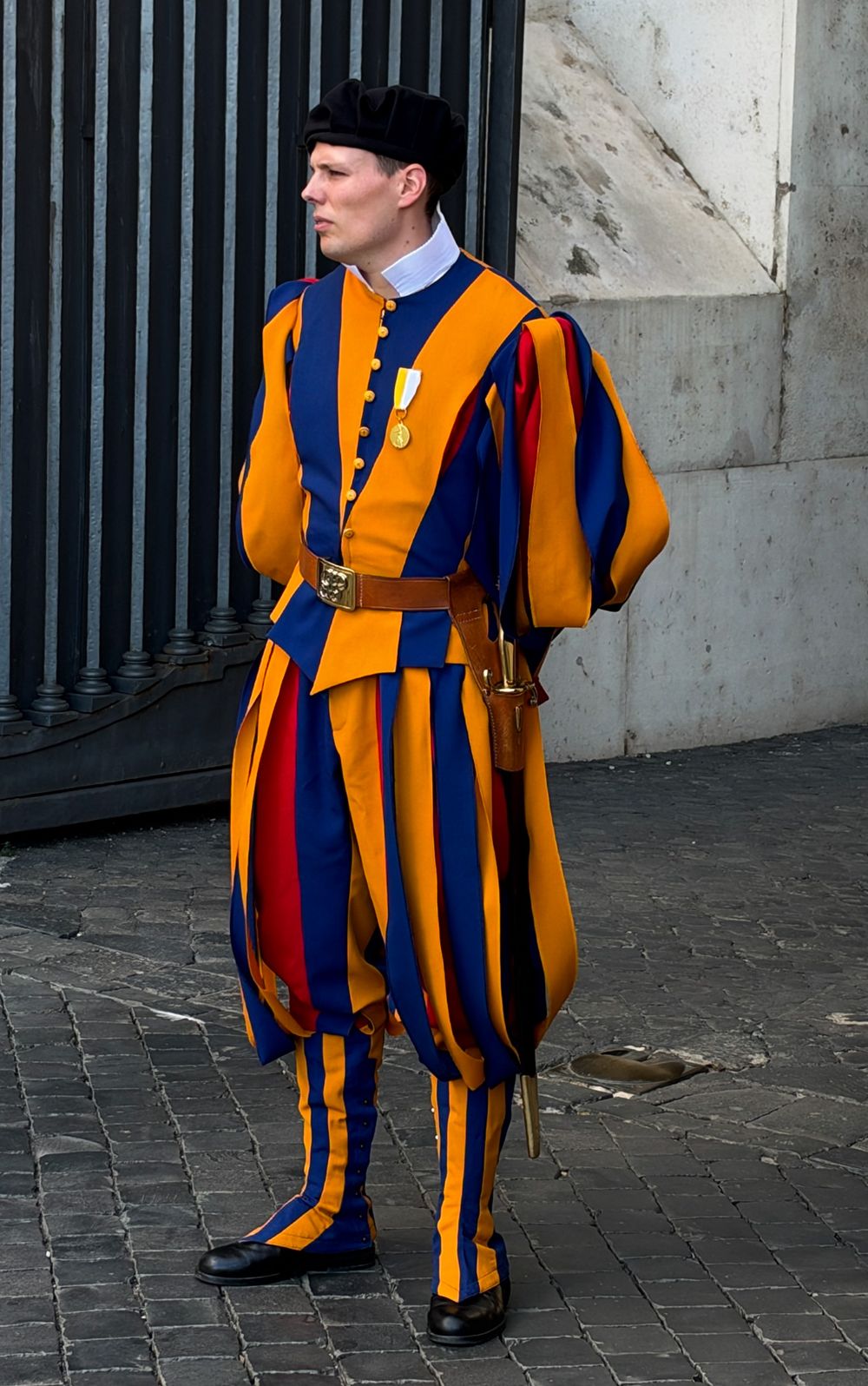 Swiss Guard in a traditional blue, red and yellow striped Renaissance uniform and black beret standing on cobblestones near an iron gate.
