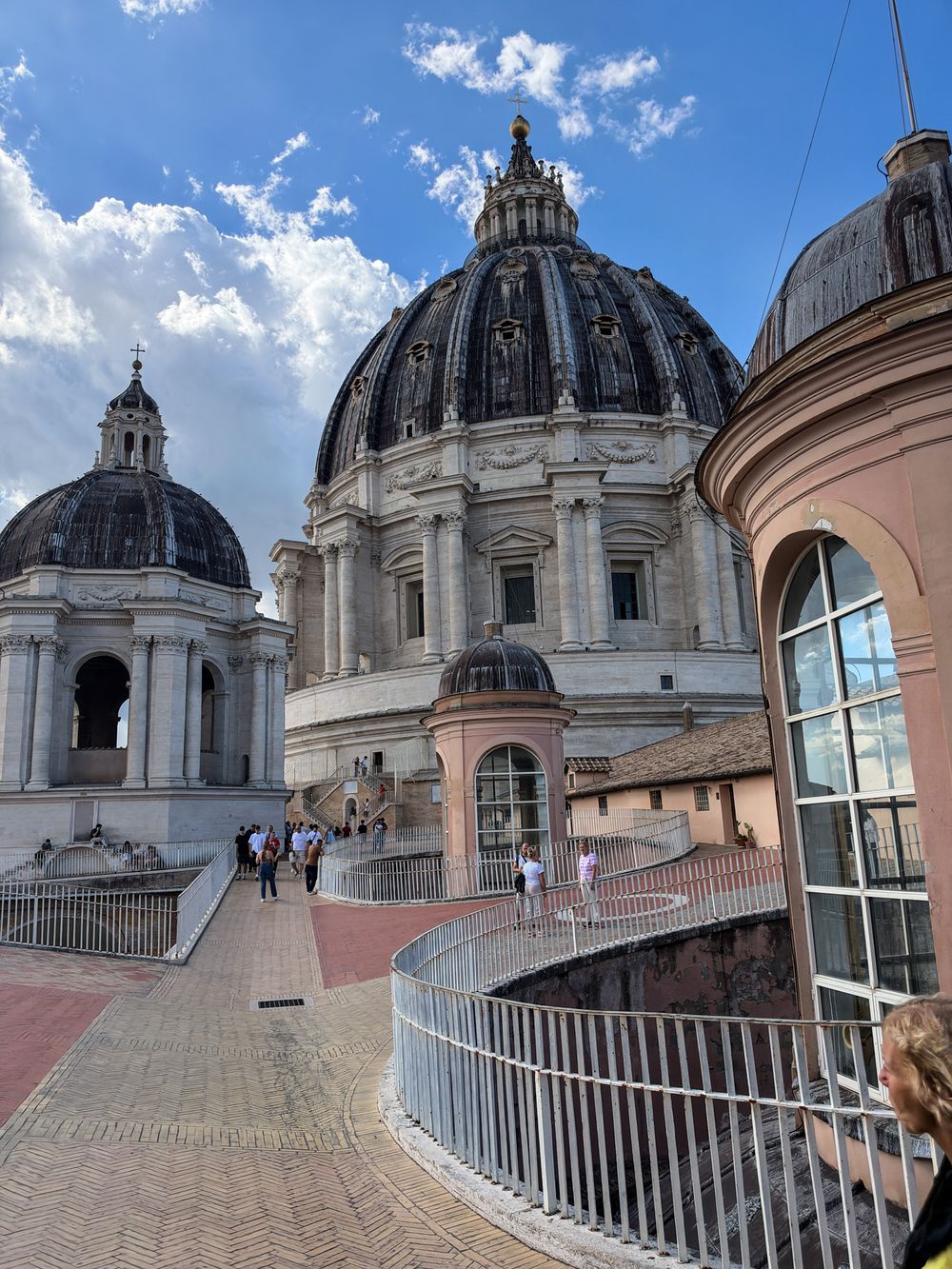 Visitors walk along a curved rooftop terrace beside the large domes of St. Peter's Basilica in Vatican City under a bright blue sky.