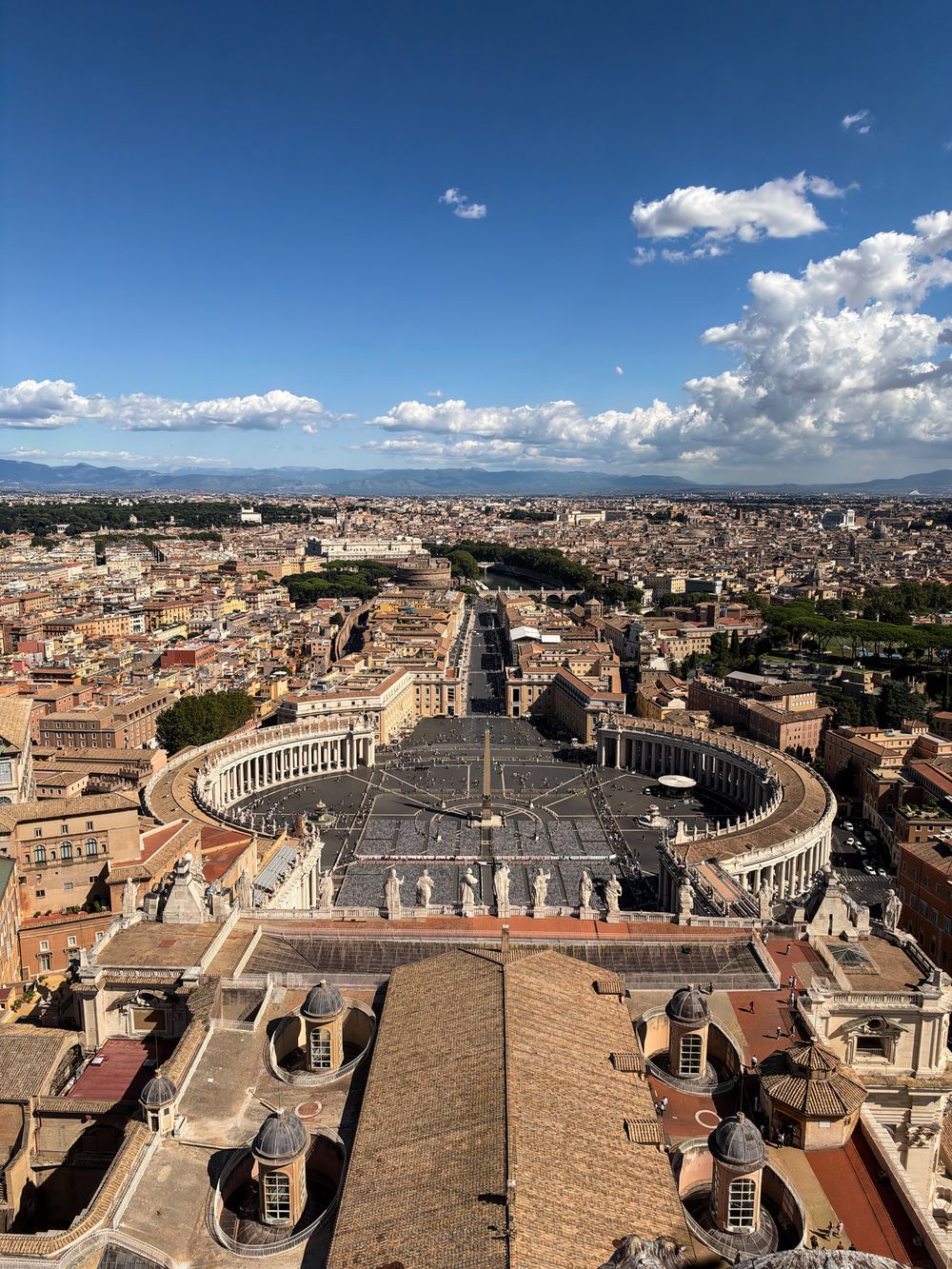 Aerial view of St. Peter's Square and its colonnades in Vatican City, seen from the basilica dome over Rome's rooftops under a blue sky.