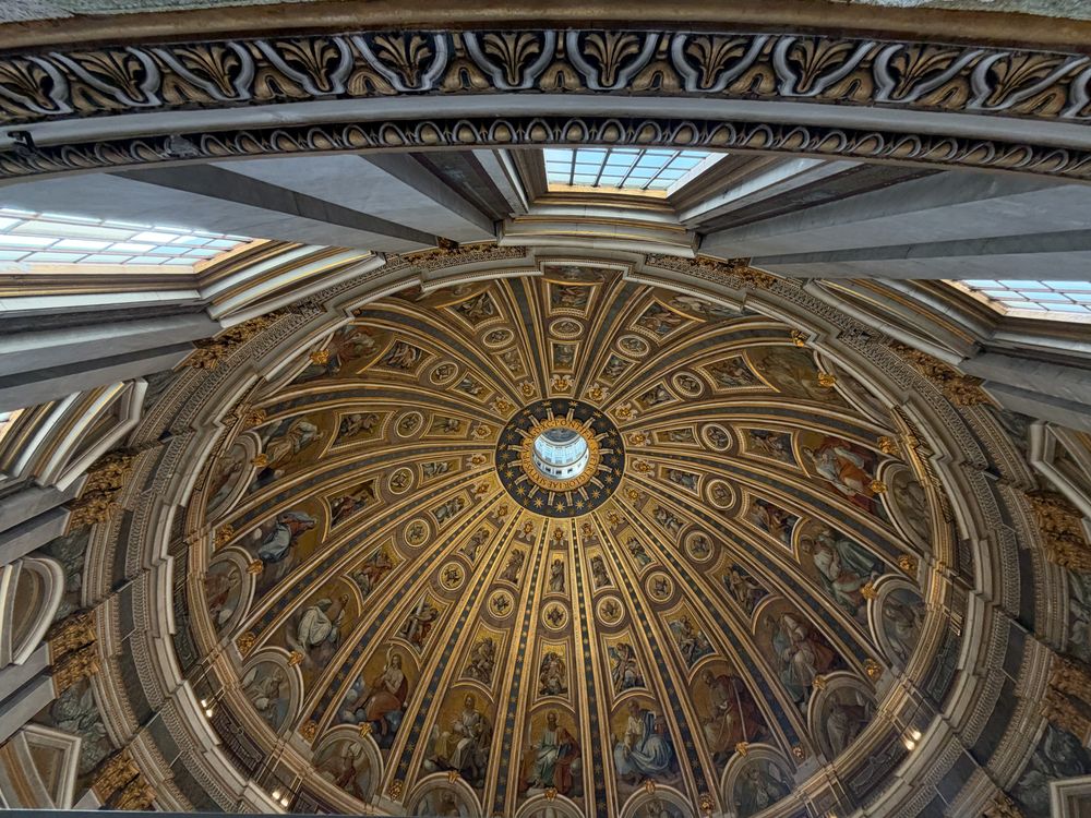 Ornate Renaissance dome interior with gilded coffered ribs, detailed mosaics, and a bright central oculus lantern, seen from below.