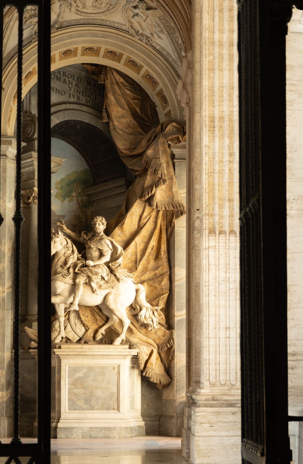 Marble equestrian statue before a sculpted golden drapery, framed by an arched marble niche and columns inside a grand baroque basilica.