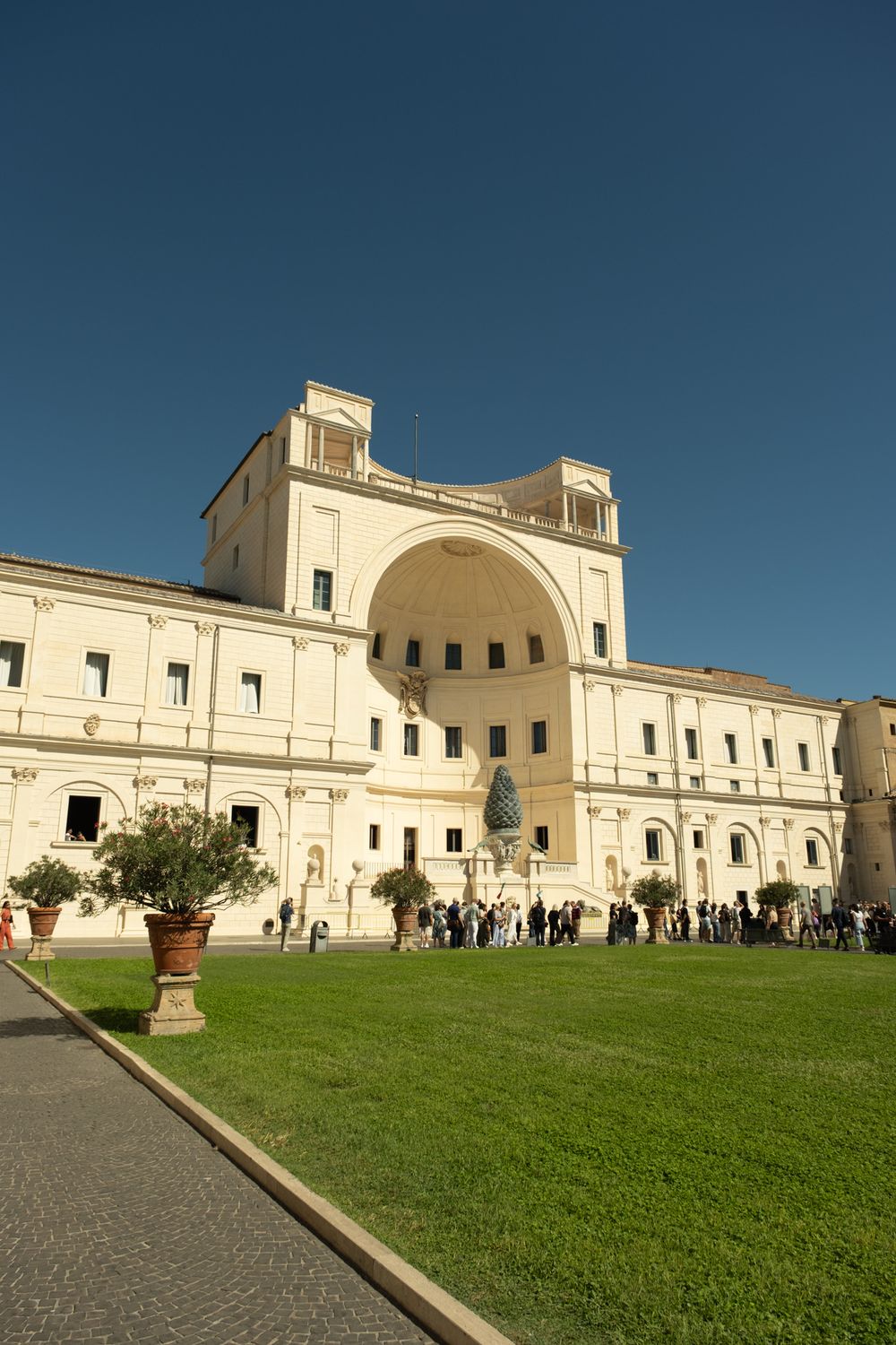 Visitors gather beside the grand Niccolo niche and giant bronze pine cone in the Vatican Museums' Pinecone Courtyard under a clear blue sky.