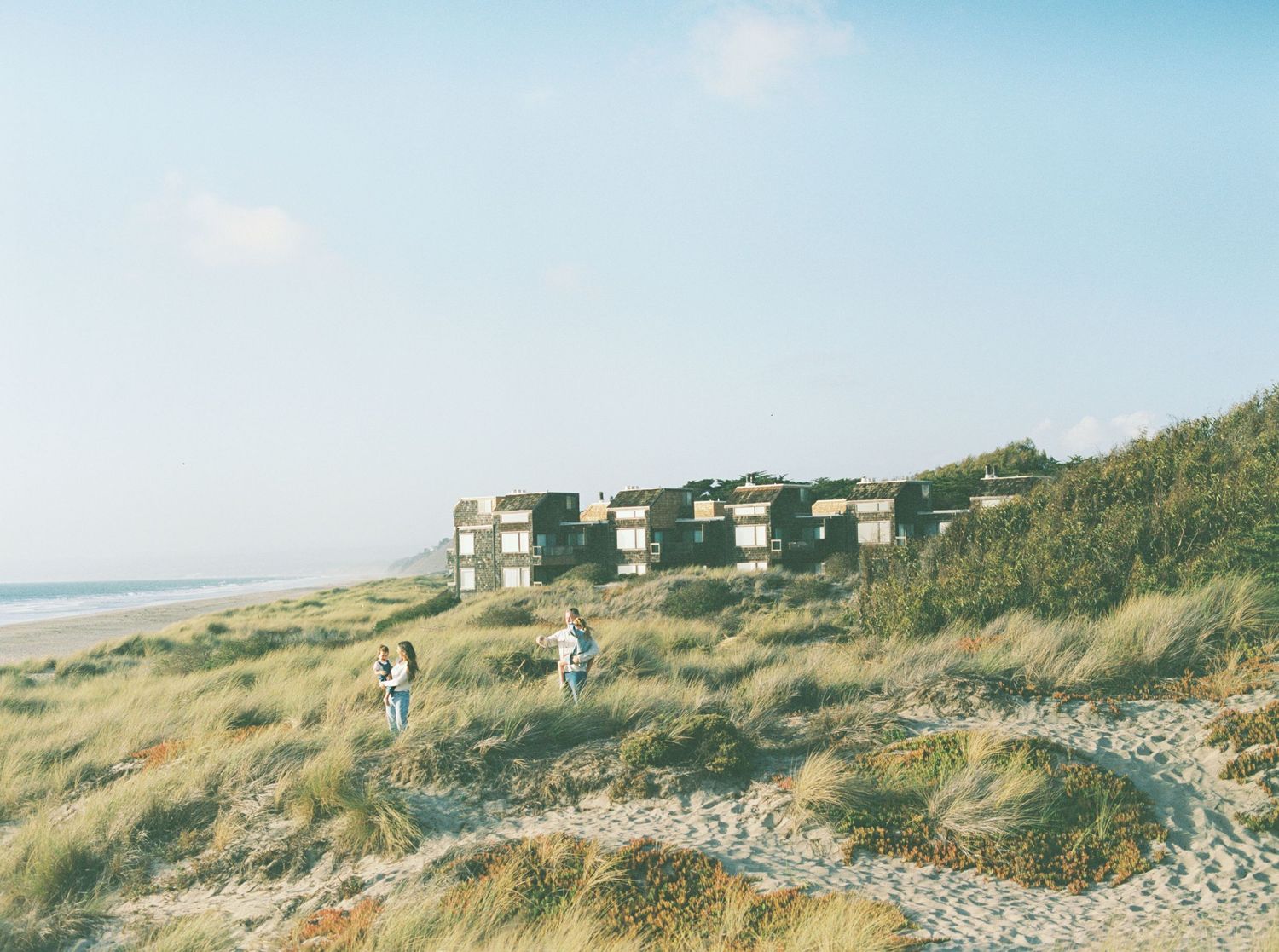 Two people walk along a sandy path through grassy coastal dunes toward a row of modern beachfront buildings overlooking the ocean.