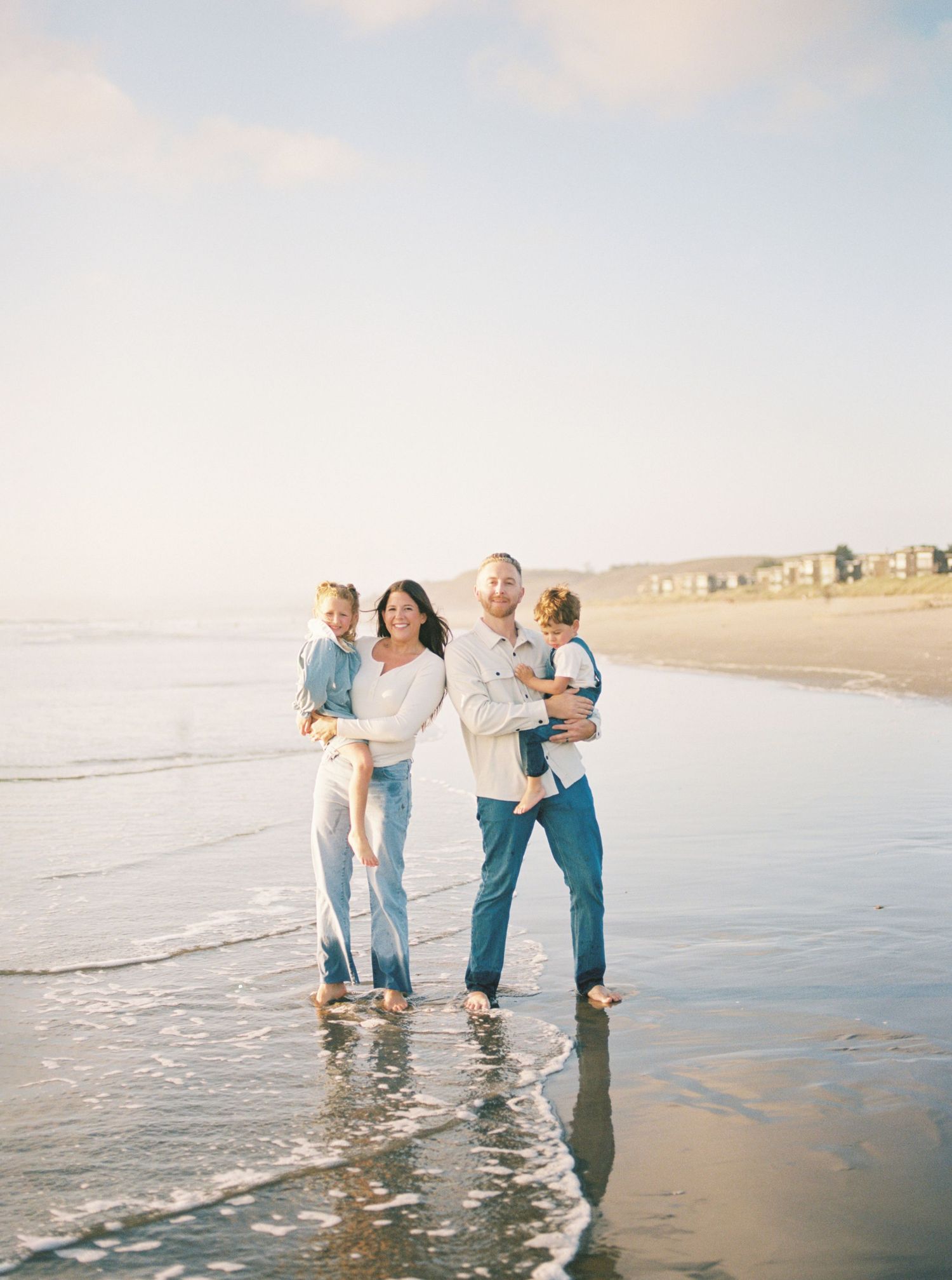 Family of four in casual white and denim standing barefoot in shallow ocean waves at the beach while holding two young children.