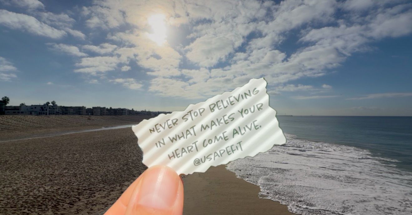 Hand holding a torn paper note with an inspirational quote against a sunny sandy beach, rolling ocean waves, and a bright blue sky.