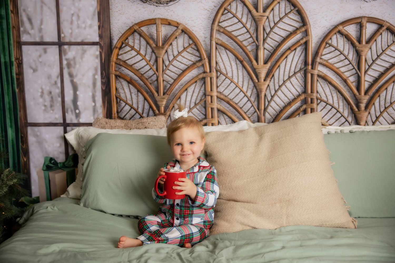 Toddler in plaid pajamas holding a red mug sits on a green bed with beige pillows in front of a woven rattan leaf headboard.