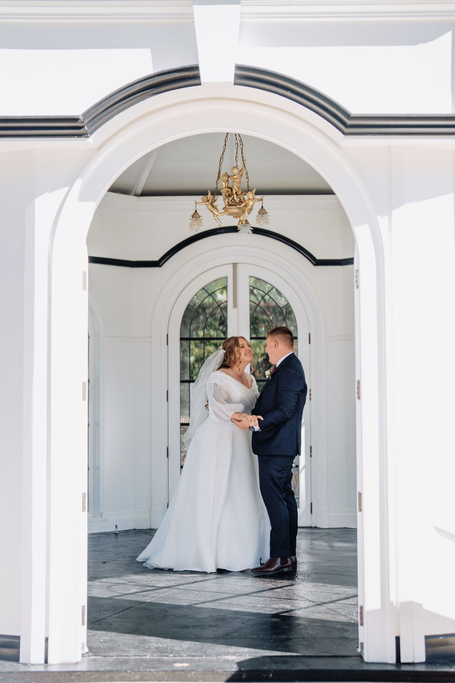 Bride in a white gown and groom in a navy suit embrace under a white archway beneath a chandelier at an elegant wedding venue.