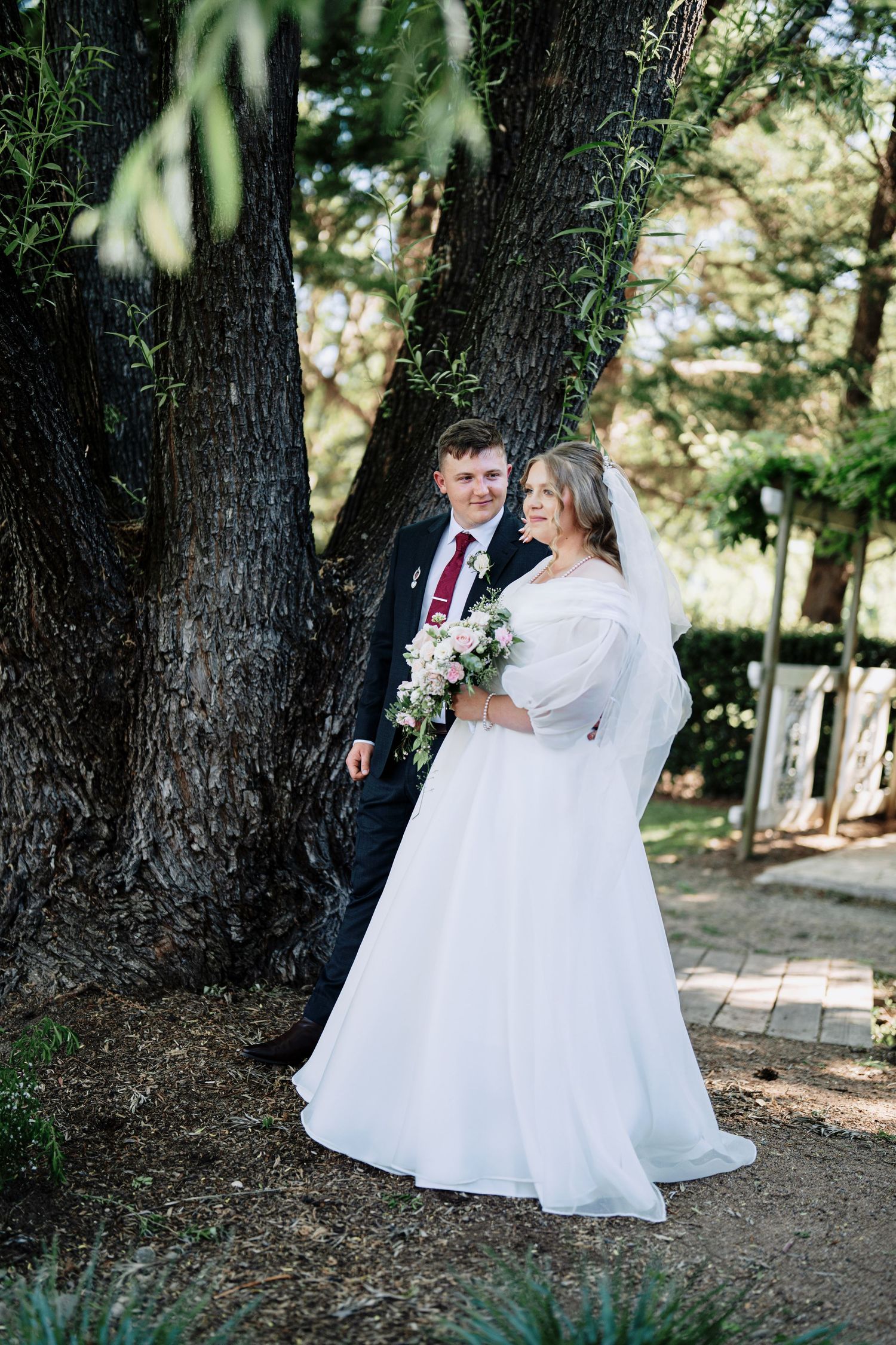 Bride in a flowing white gown and veil holding a pink floral bouquet stands beside a groom in a navy suit beneath a large tree.