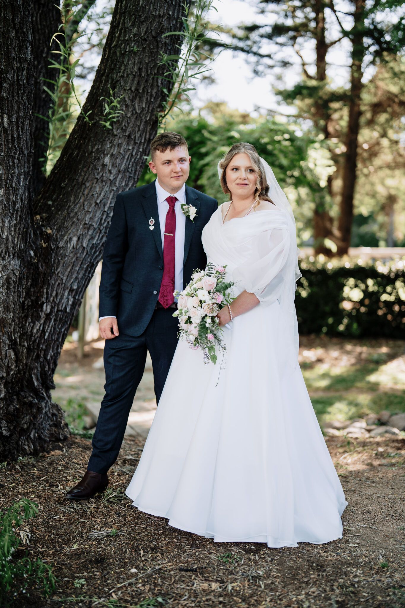 Bride in a white gown with veil holding a bouquet stands beside a groom in a navy suit and burgundy tie beneath a large tree.
