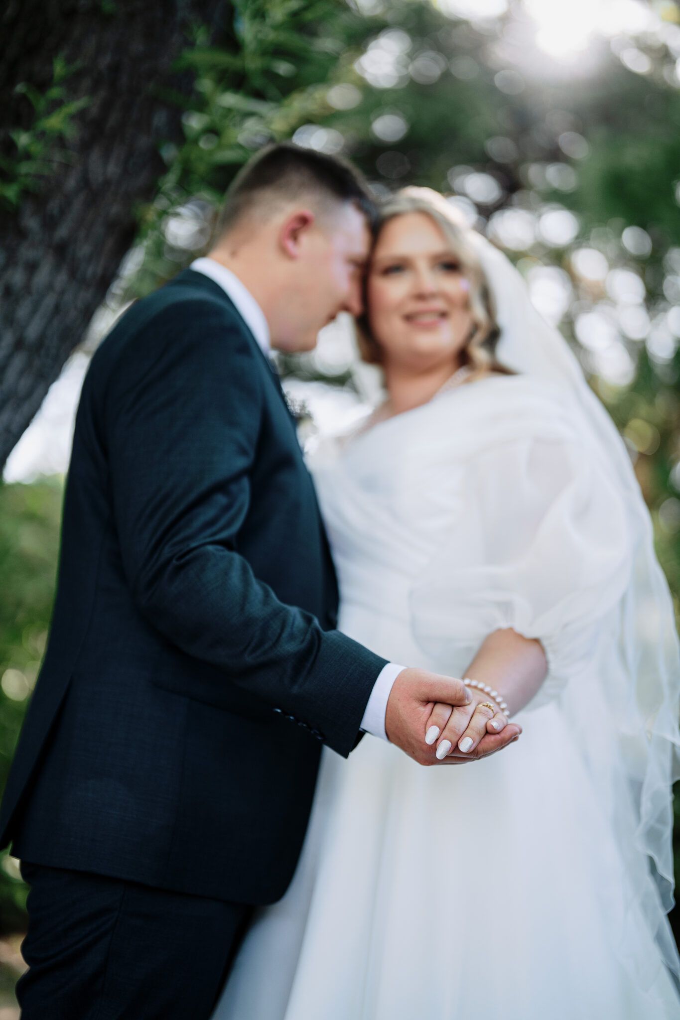 Bride in a white tulle gown and veil and groom in a dark suit hold hands, showing the wedding ring, beneath sunlit trees.