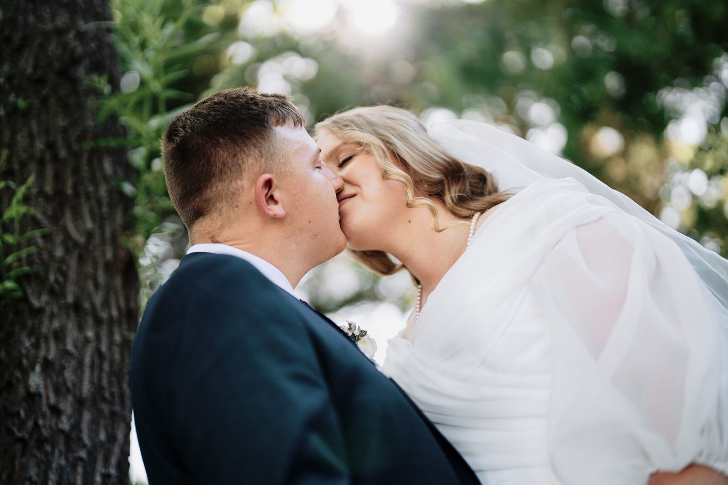 Groom in a dark suit kisses a smiling bride in a flowing white veil outdoors beneath sunlit green trees on their wedding day.