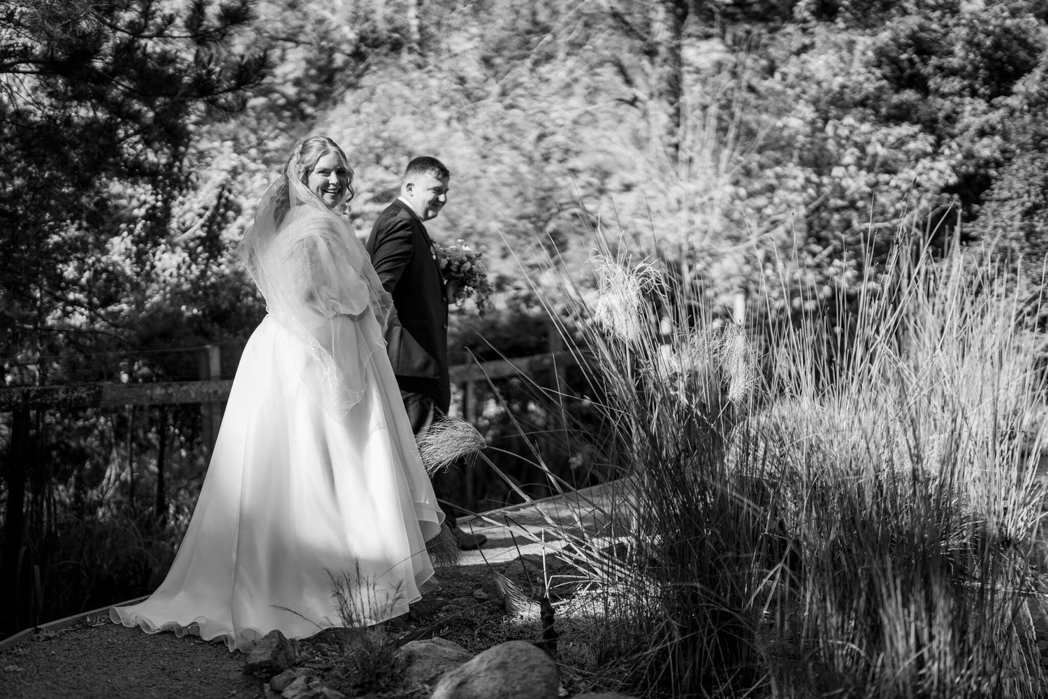 Black and white wedding photo of a bride in a long gown and veil walking with her groom along a garden path beside tall grasses.