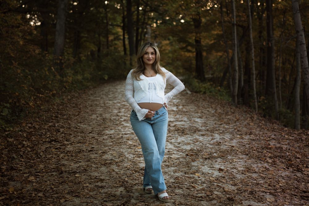 Woman in a white long-sleeve top and blue jeans stands on a leaf-covered forest trail surrounded by autumn trees.