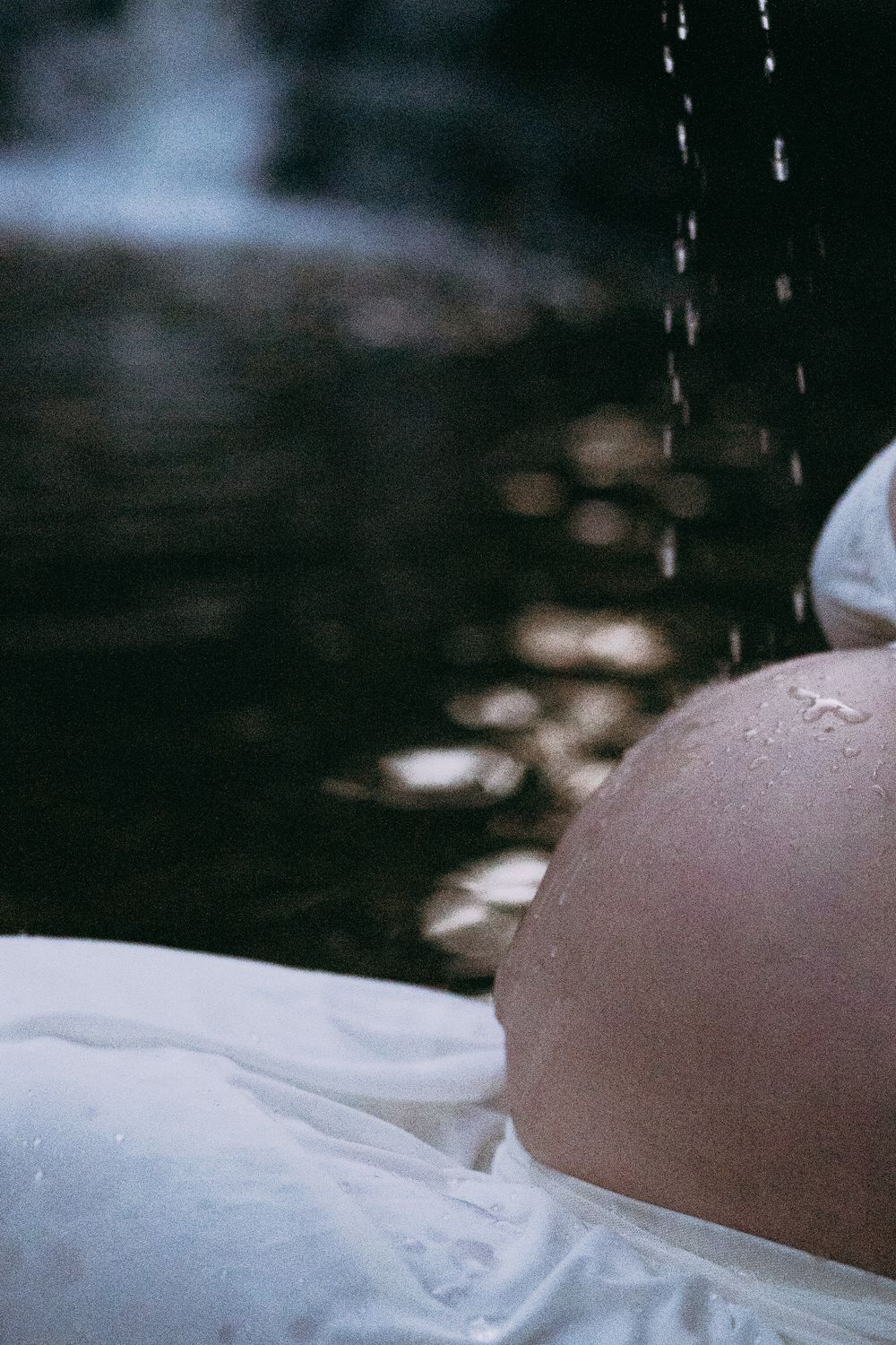 Water droplets fall onto a pregnant belly draped in white fabric, with soft light reflecting on a dark stream in the background In Lemont