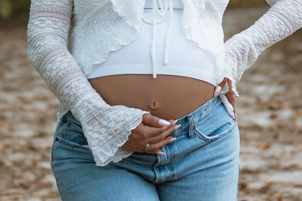 Pregnant woman in a white crop top and lace sleeves gently cradles her bare baby bump while wearing light blue jeans outdoors.