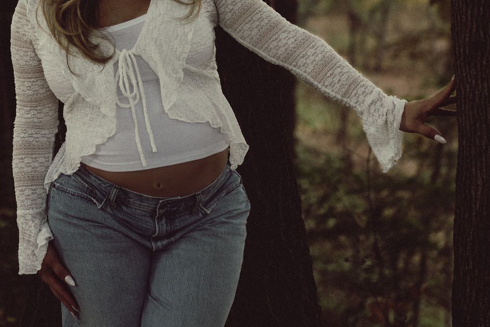 Woman in a white ruffled lace cardigan, tied crop top and light blue jeans leaning against a tree trunk in a wooded forest setting.