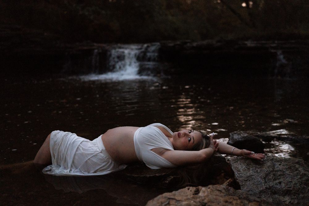 Pregnant woman in a white crop top and flowing skirt reclines on rocks beside a still forest pool with a small waterfall behind her.