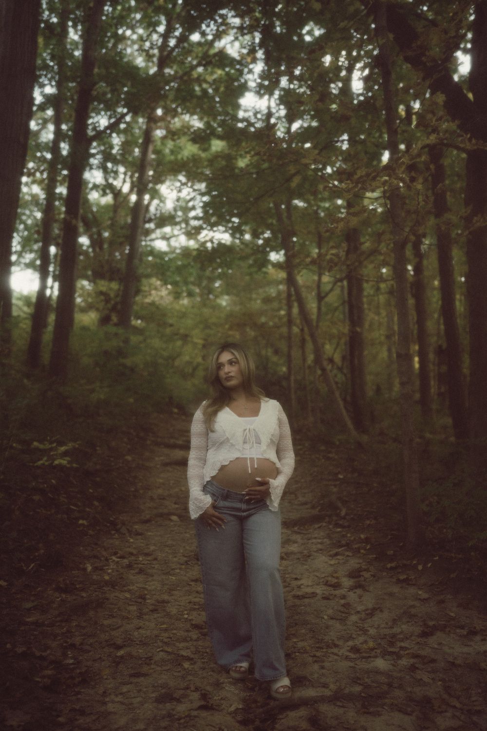 Woman in a white lace top and gray wide-leg jeans stands on a dirt path in a sunlit wooded forest, resting her hand on her belly.