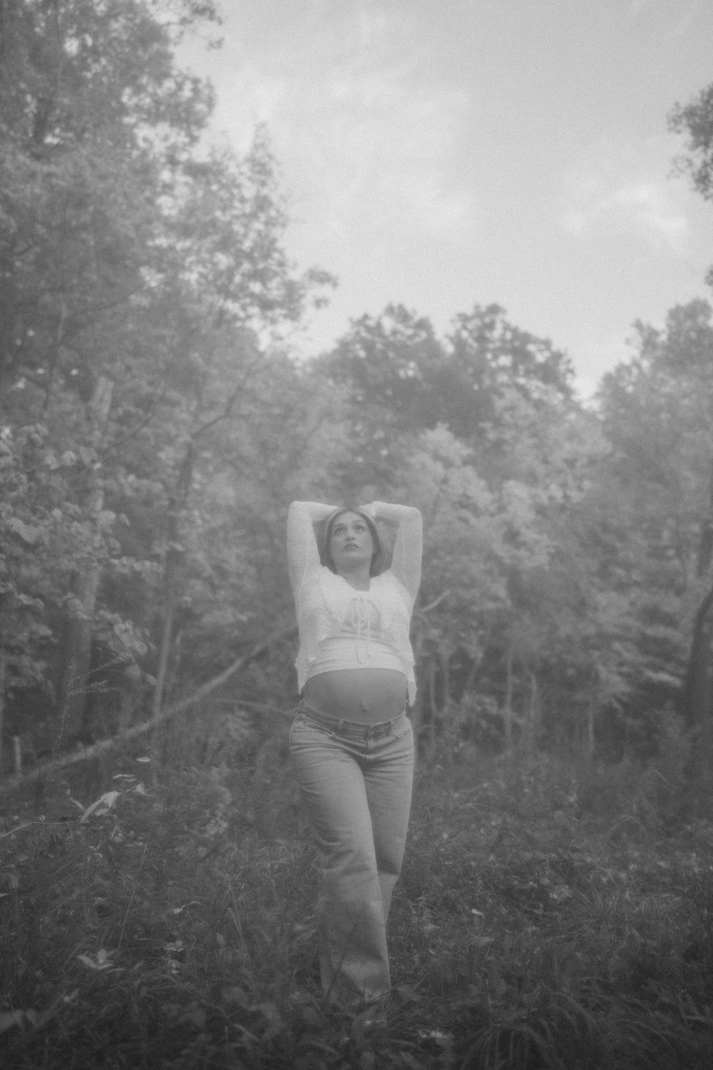 Black and white photo of a woman with arms raised overhead, standing in a misty forest clearing surrounded by tall trees.