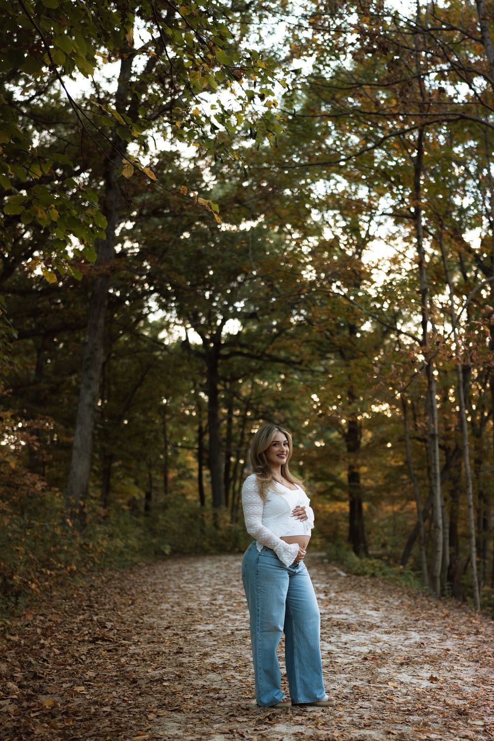 Pregnant woman in white top and wide-leg jeans cradling her baby bump on a leaf-covered forest path in autumn.