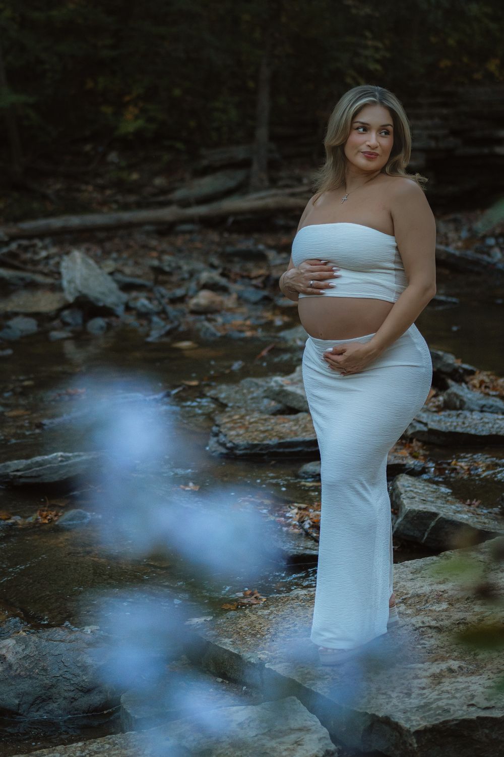 Pregnant woman in a white crop top and long white skirt cradles her baby bump while posing on rocks beside a woodland creek.