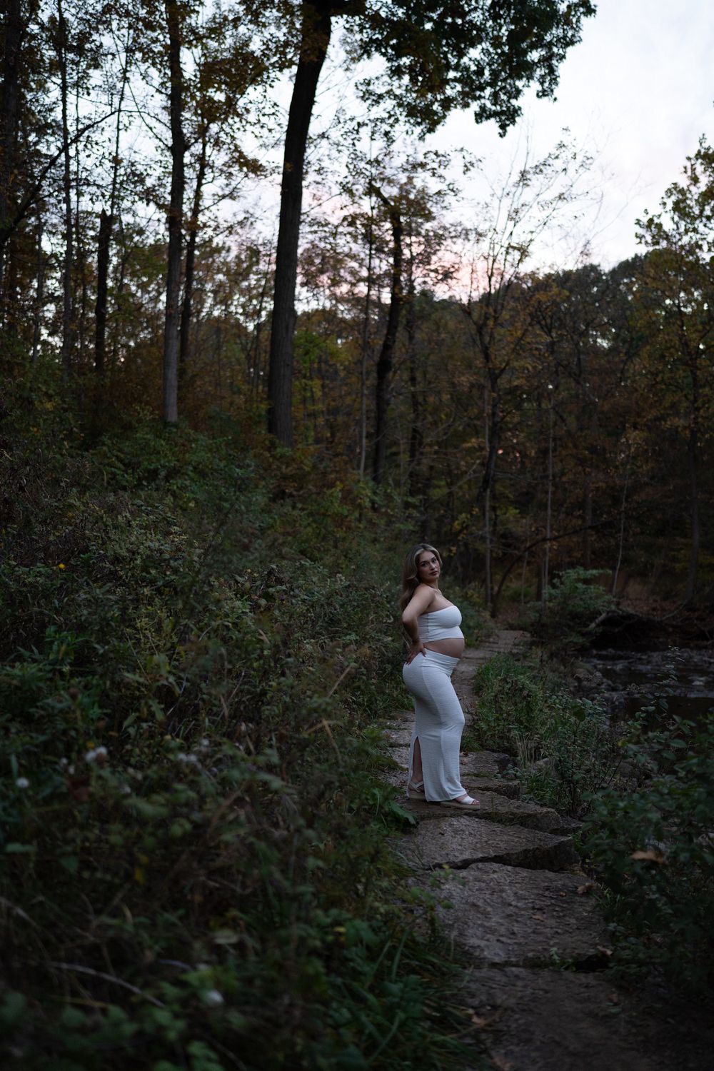 Pregnant woman in a white fitted two-piece outfit posing on a stone path in a woodland setting at dusk for a maternity photoshoot.