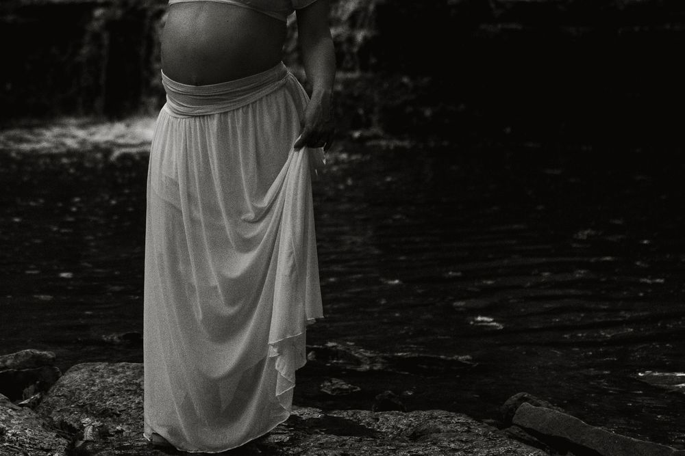 Black and white photo of a pregnant woman in a flowing white skirt standing on rocks beside a stream, with her baby bump bare.