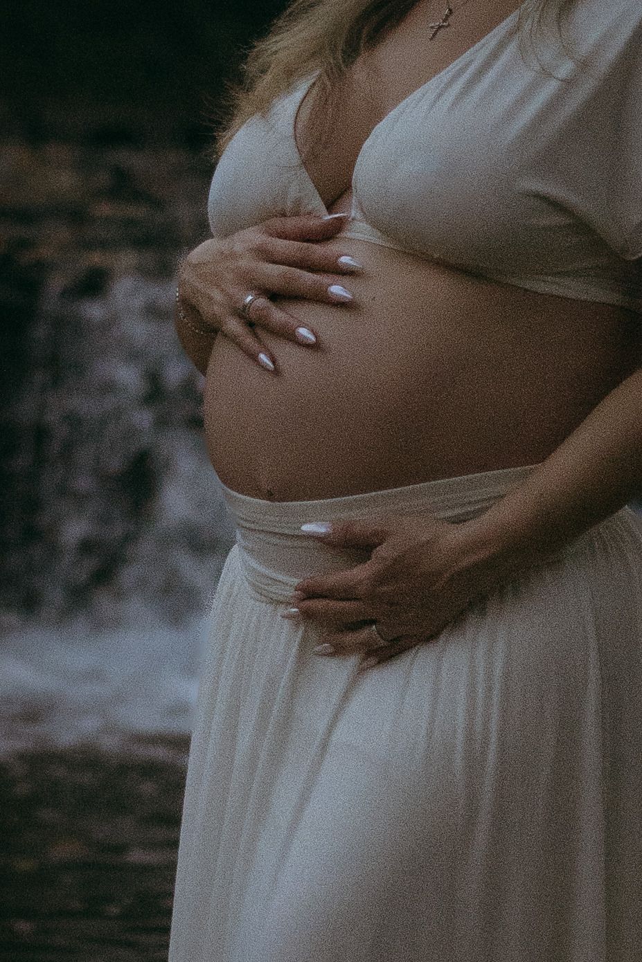 Pregnant woman in a flowing cream skirt and crop top cradles her bare baby bump beside a waterfall in a moody maternity photo.