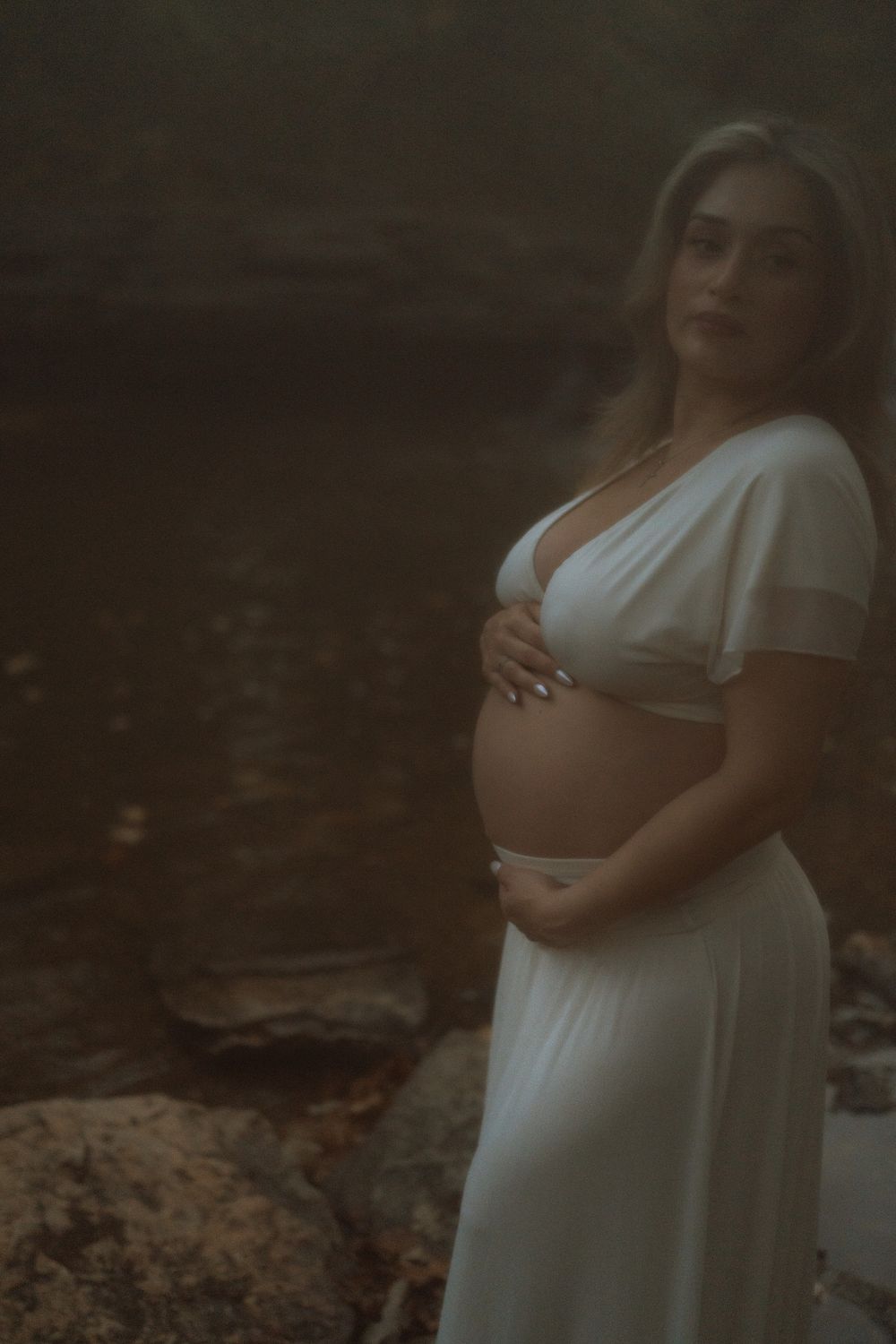 Pregnant woman in a white two-piece outfit cradles her belly while standing on rocks beside a misty, moody river at dusk.