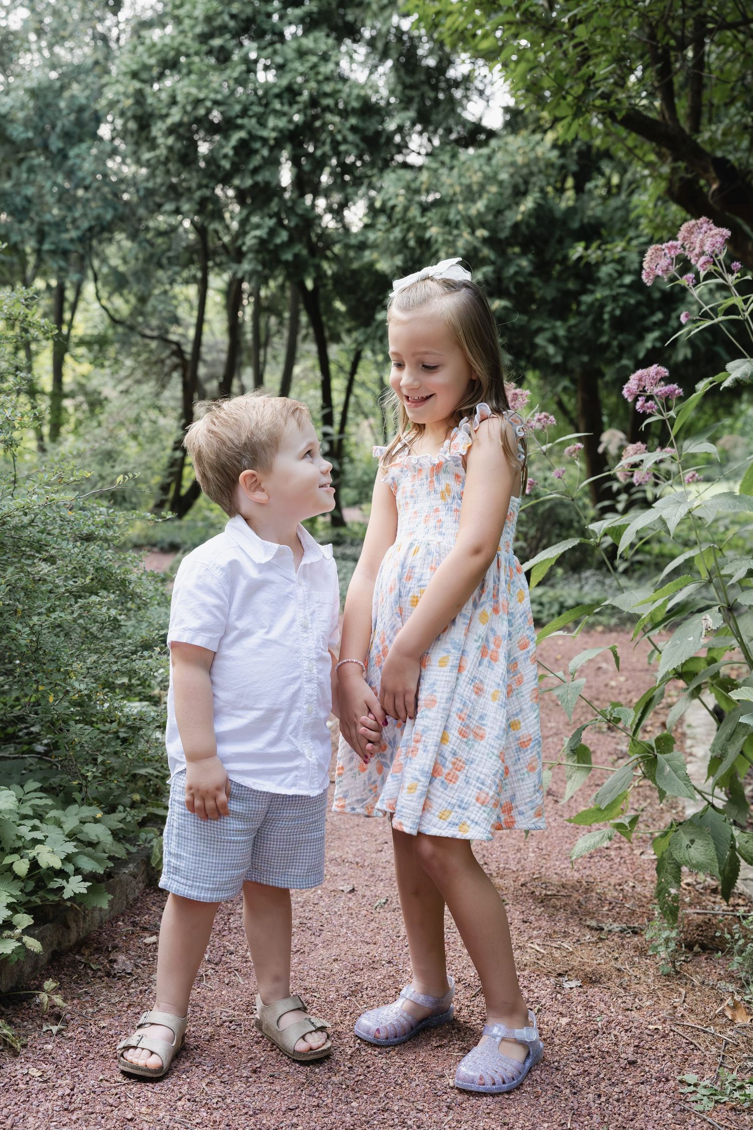 Two young children in formal attire hold hands on a garden path surrounded by lush green trees.