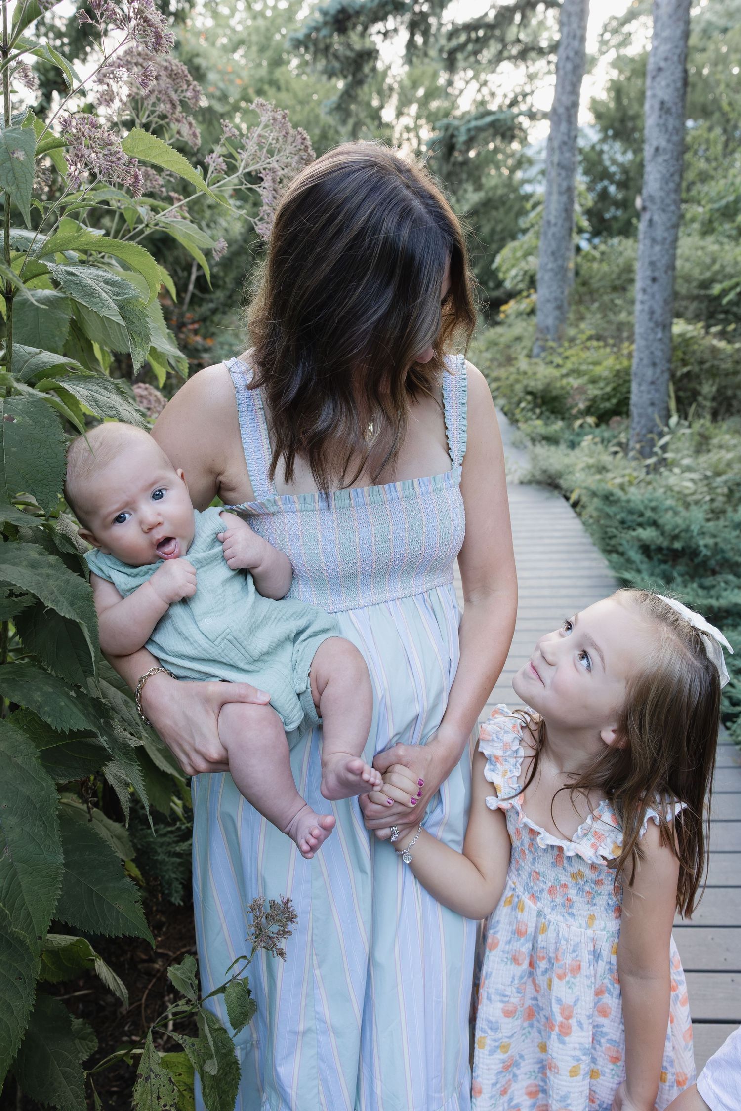 Mother in blue striped dress holds infant while young daughter in floral dress looks up at her outdoors.
