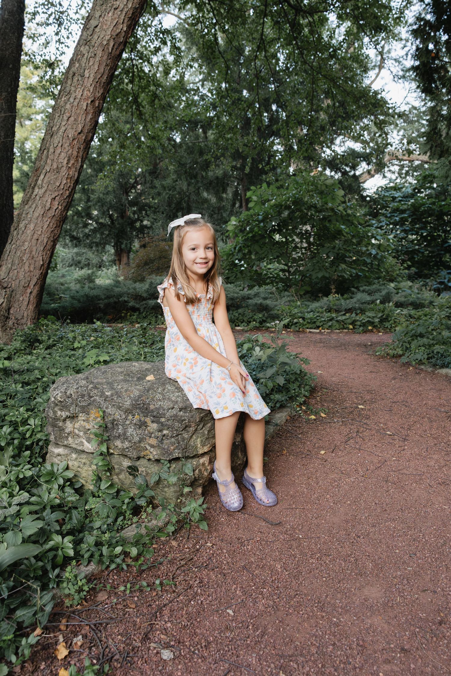 Young girl in floral dress sitting on stone wall along garden path, smiling among lush green trees.