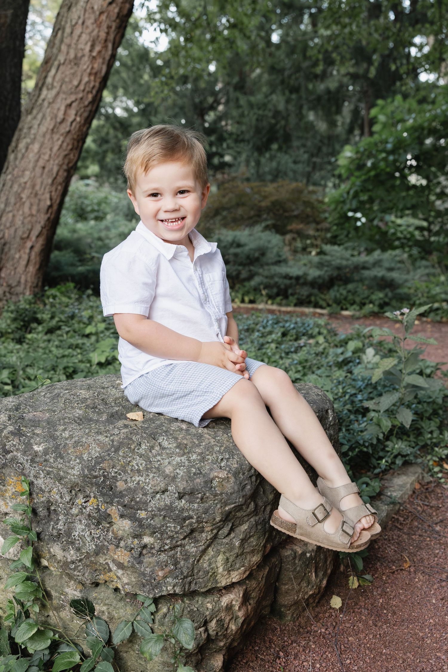 Happy toddler boy in white outfit sitting on large rock in lush green park setting.