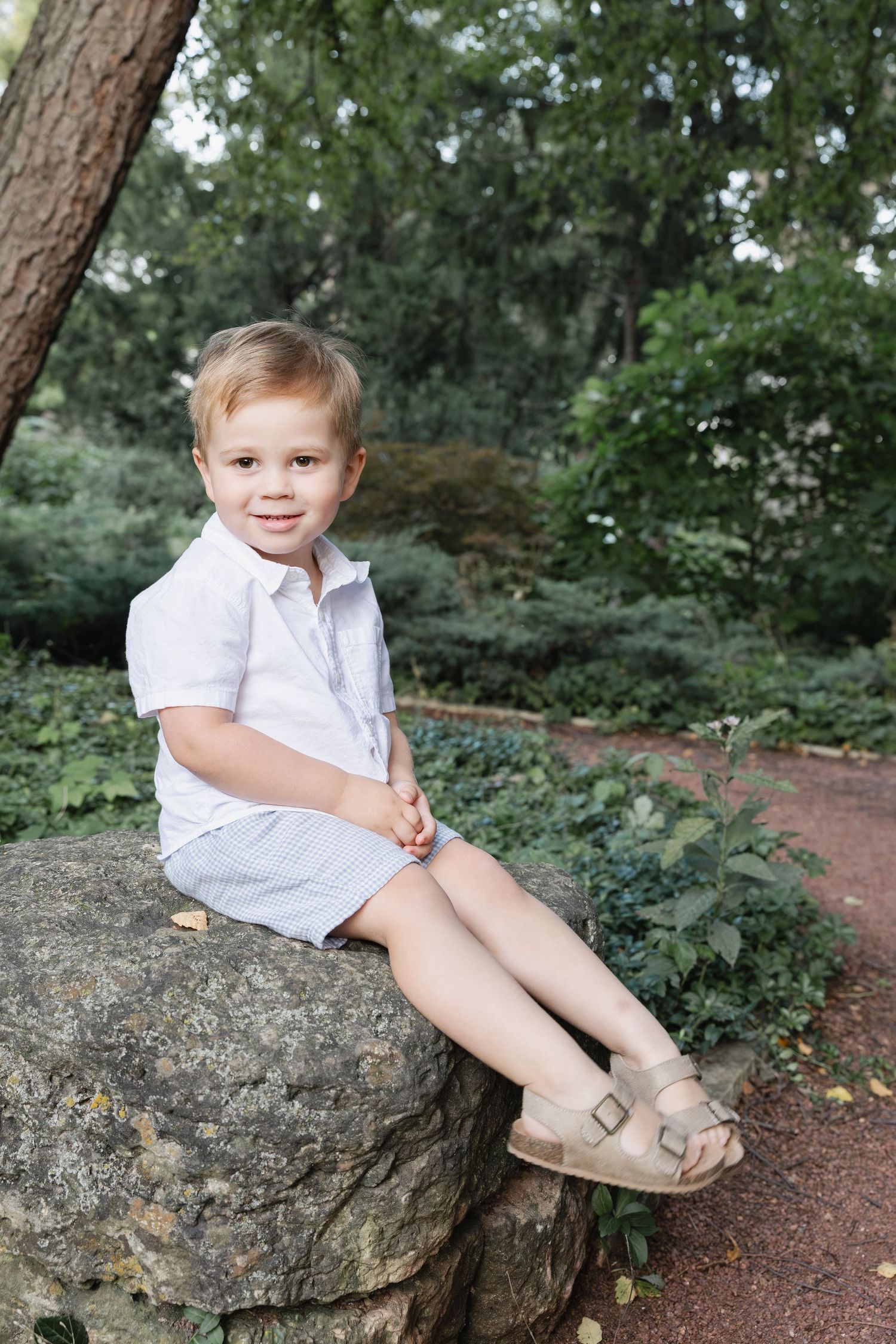 Adorable young boy in white outfit sits smiling on a large rock in a lush green garden setting.