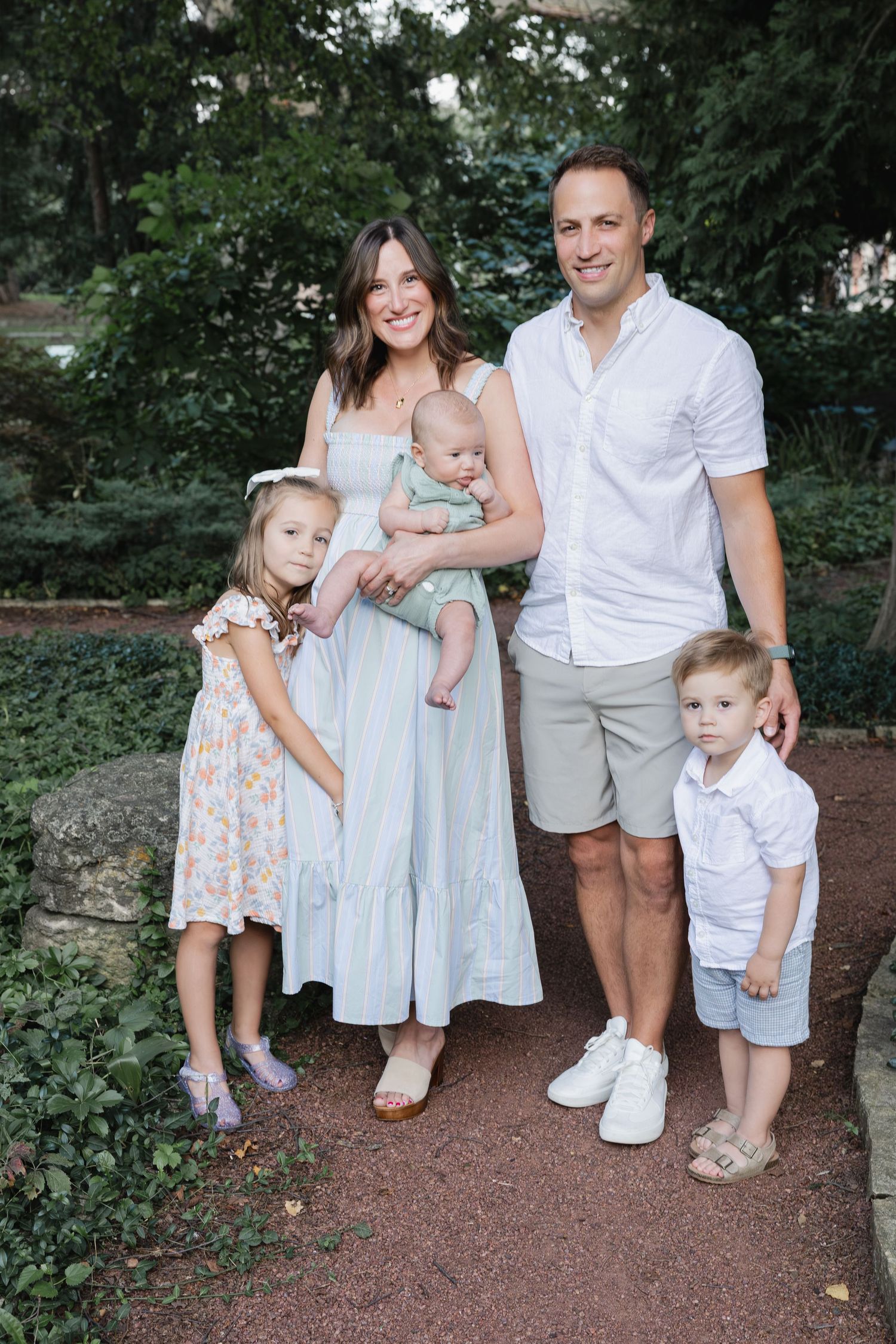 A happy family of five poses outdoors, with parents holding a baby and two young children standing beside them.