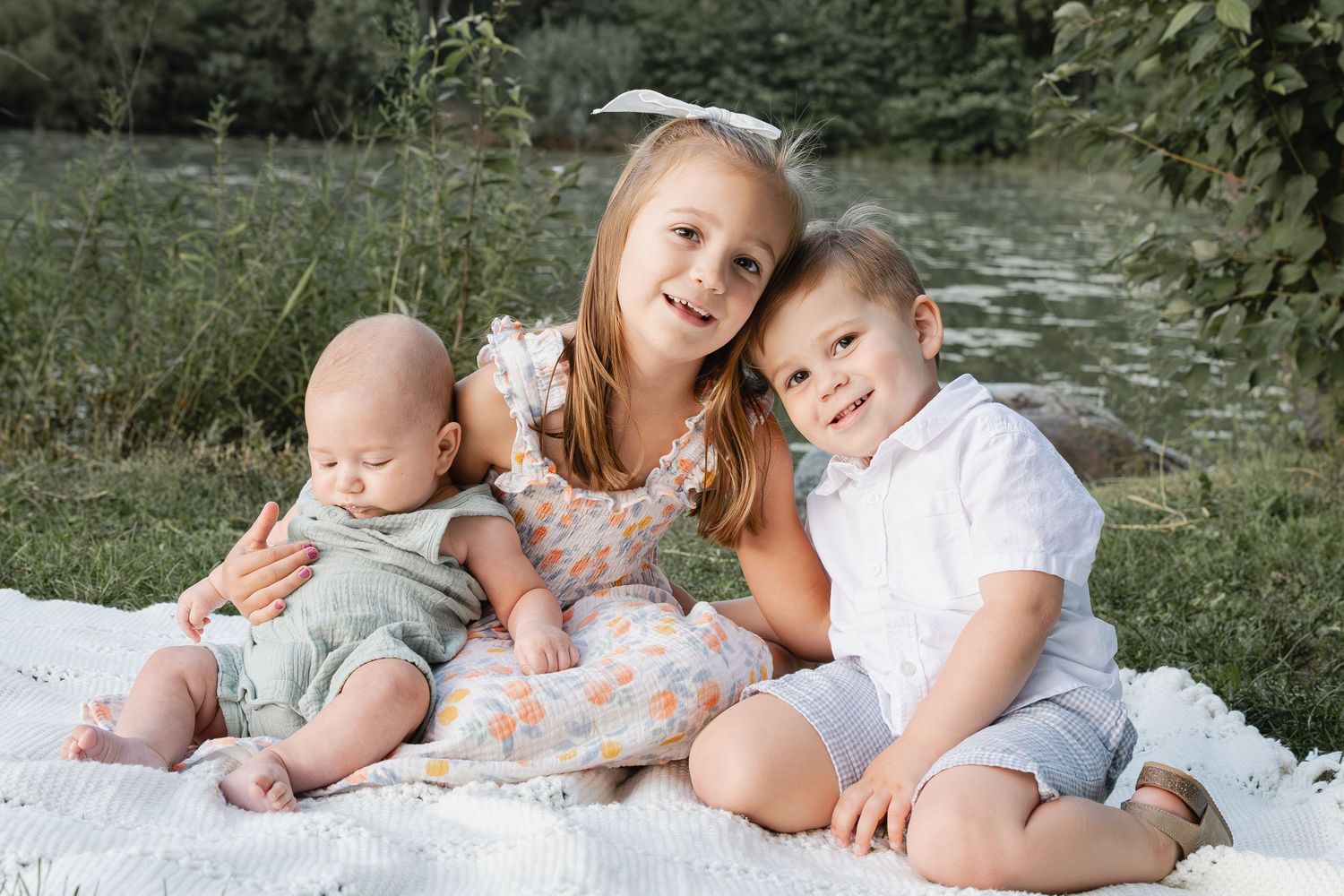 Three young siblings pose together on white blanket outdoors near greenery for family portrait.