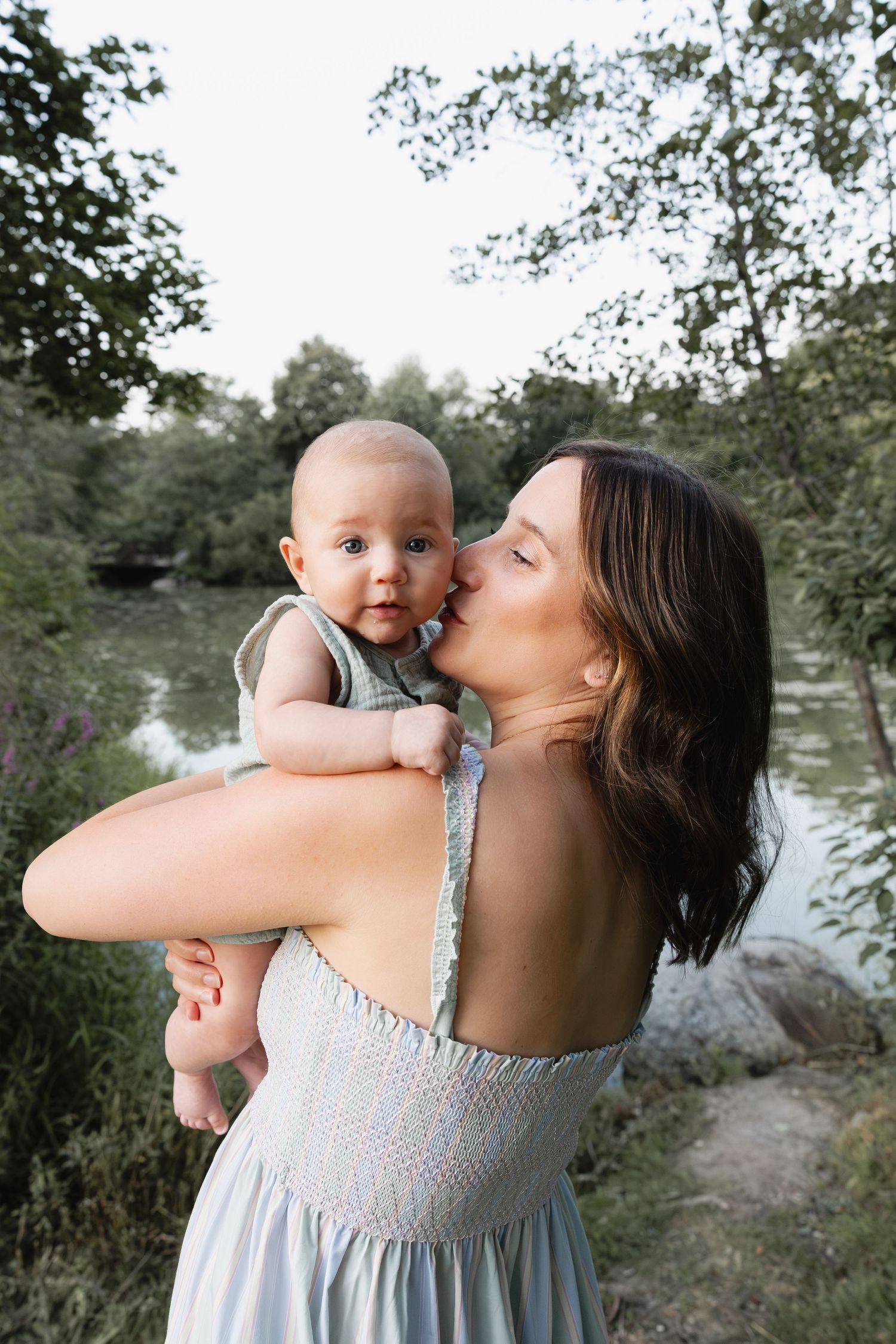 A mother in a light blue dress kisses her baby outdoors near a serene river surrounded by lush green trees.