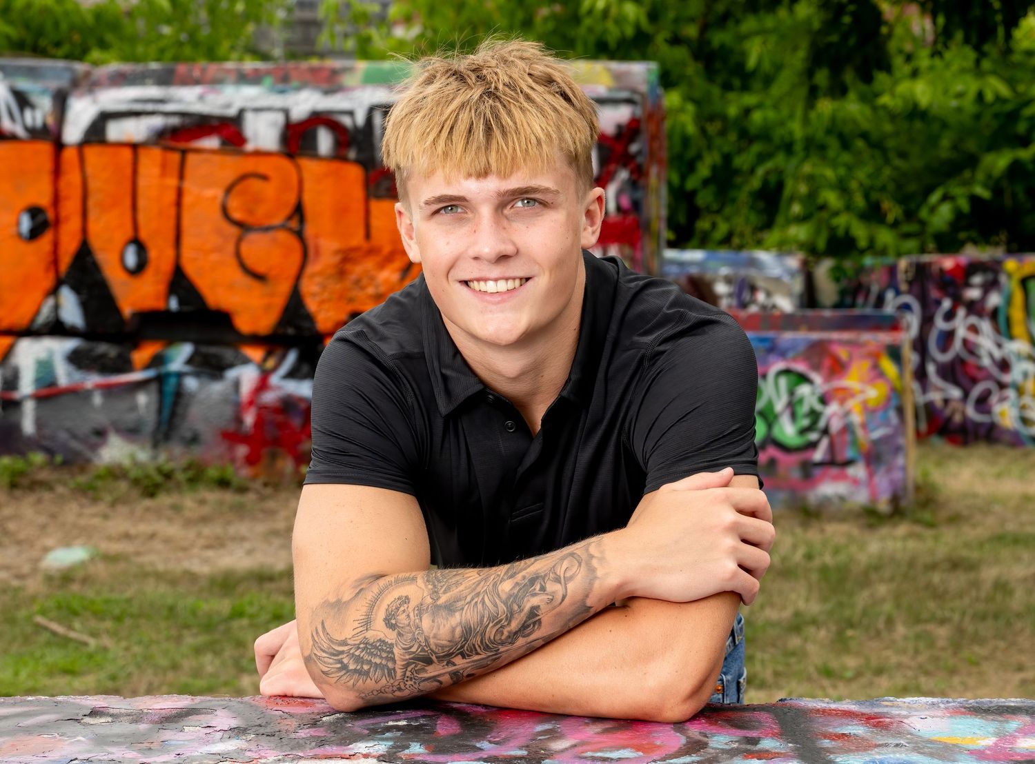 Young man with tattoos and blonde hair smiling, leaning on graffiti wall outdoors.