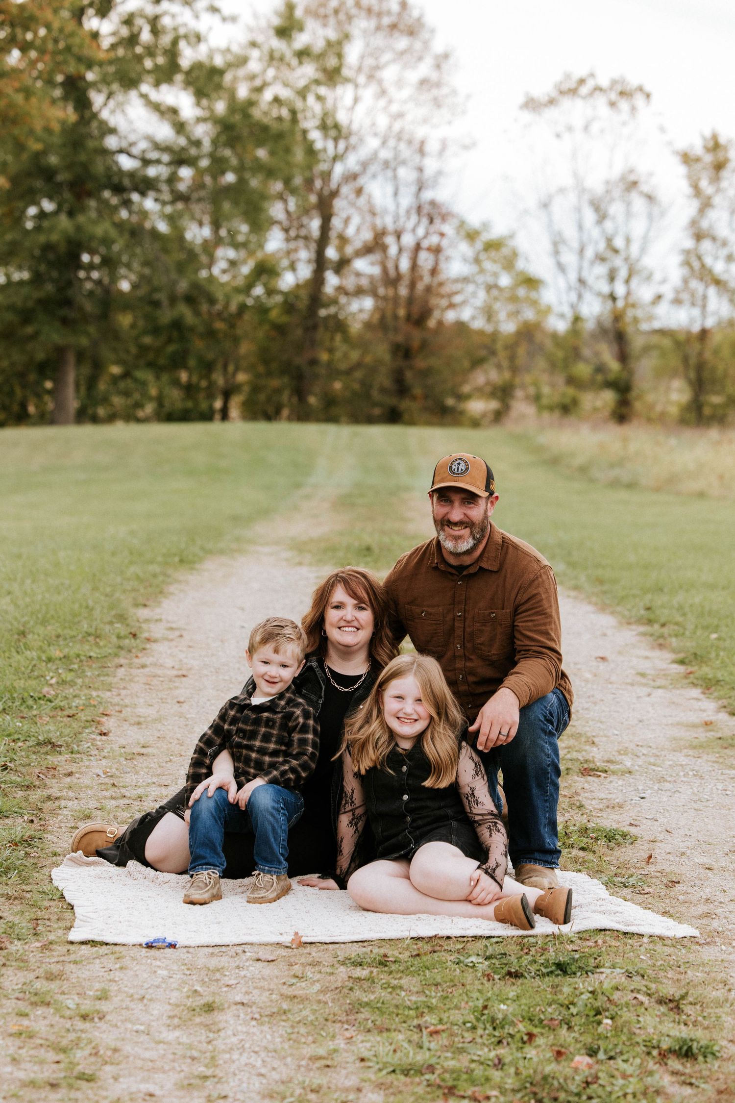 Family of four posing on a blanket along a scenic gravel path surrounded by autumn trees during an outdoor photo session.