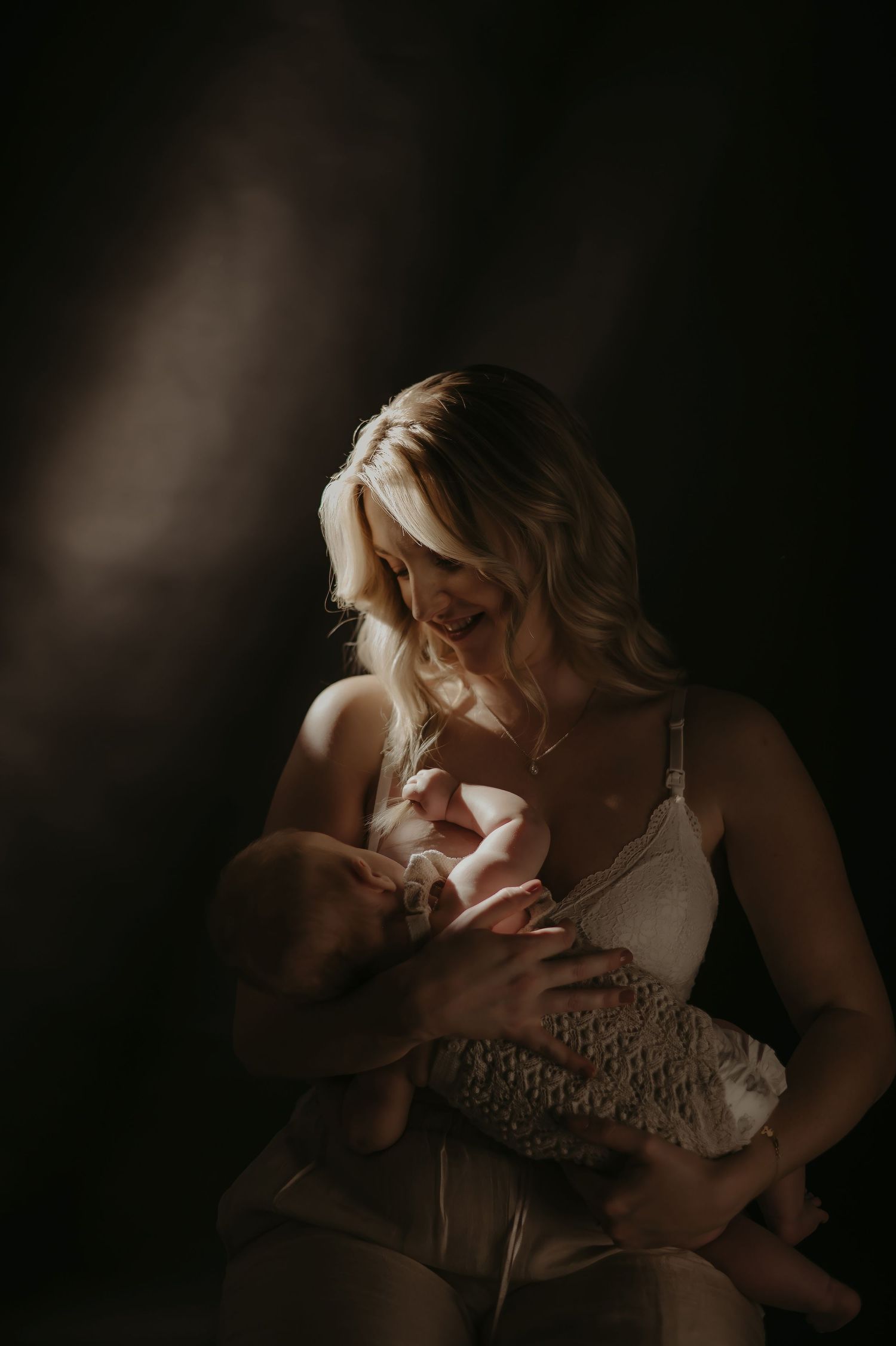 A blonde mother tenderly holds her newborn baby in soft, dramatic lighting against a dark background.