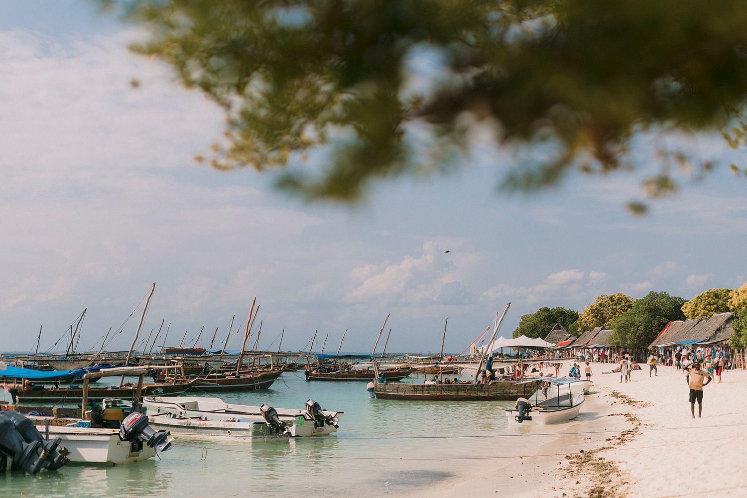 Wooden boats moored along a sandy tropical beach with clear turquoise water and visitors walking the shoreline.